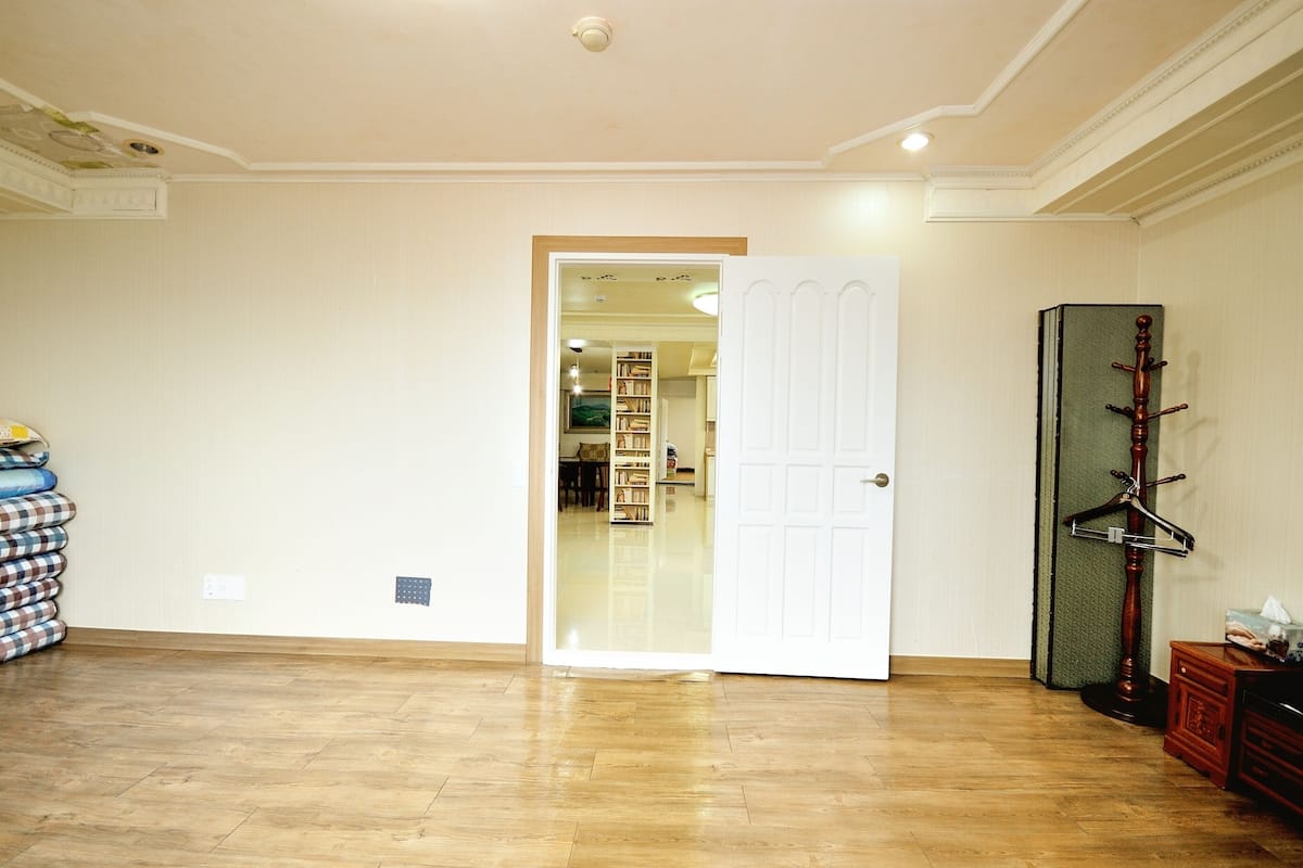 A room with a wooden floor, a white door, a coat rack, and a stack of folded blankets.
