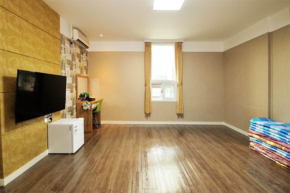 A room with a flat-screen TV, a small refrigerator, a dining table with chairs, a window with curtains, and a stack of folded blankets.