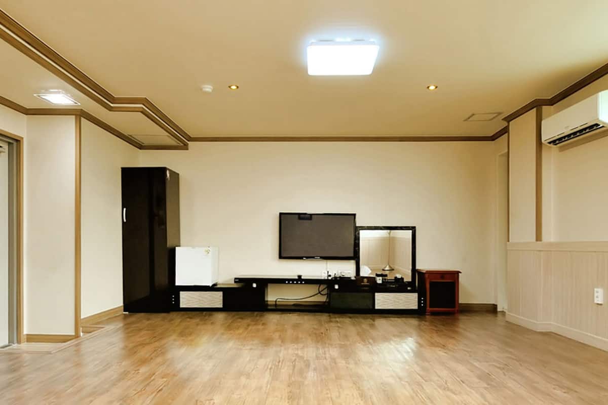 A room with a wooden floor, a flat-screen TV mounted on the wall, a desk with a computer, and a small table with a lamp.