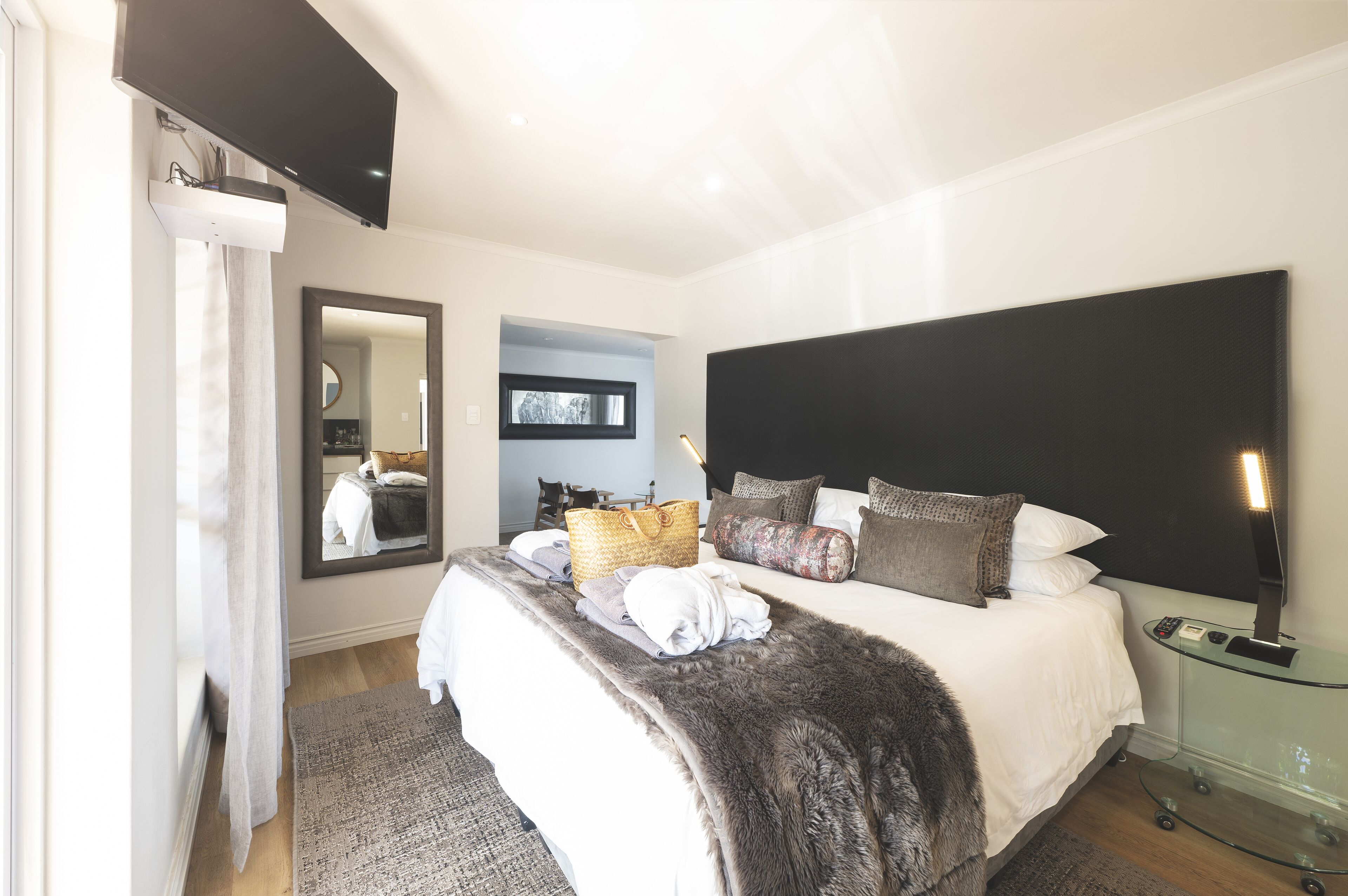 Un dormitorio moderno con una cama grande, un televisor de pantalla plana montado en la pared, una mesita de noche con lámpara y un espejo sobre la cama.