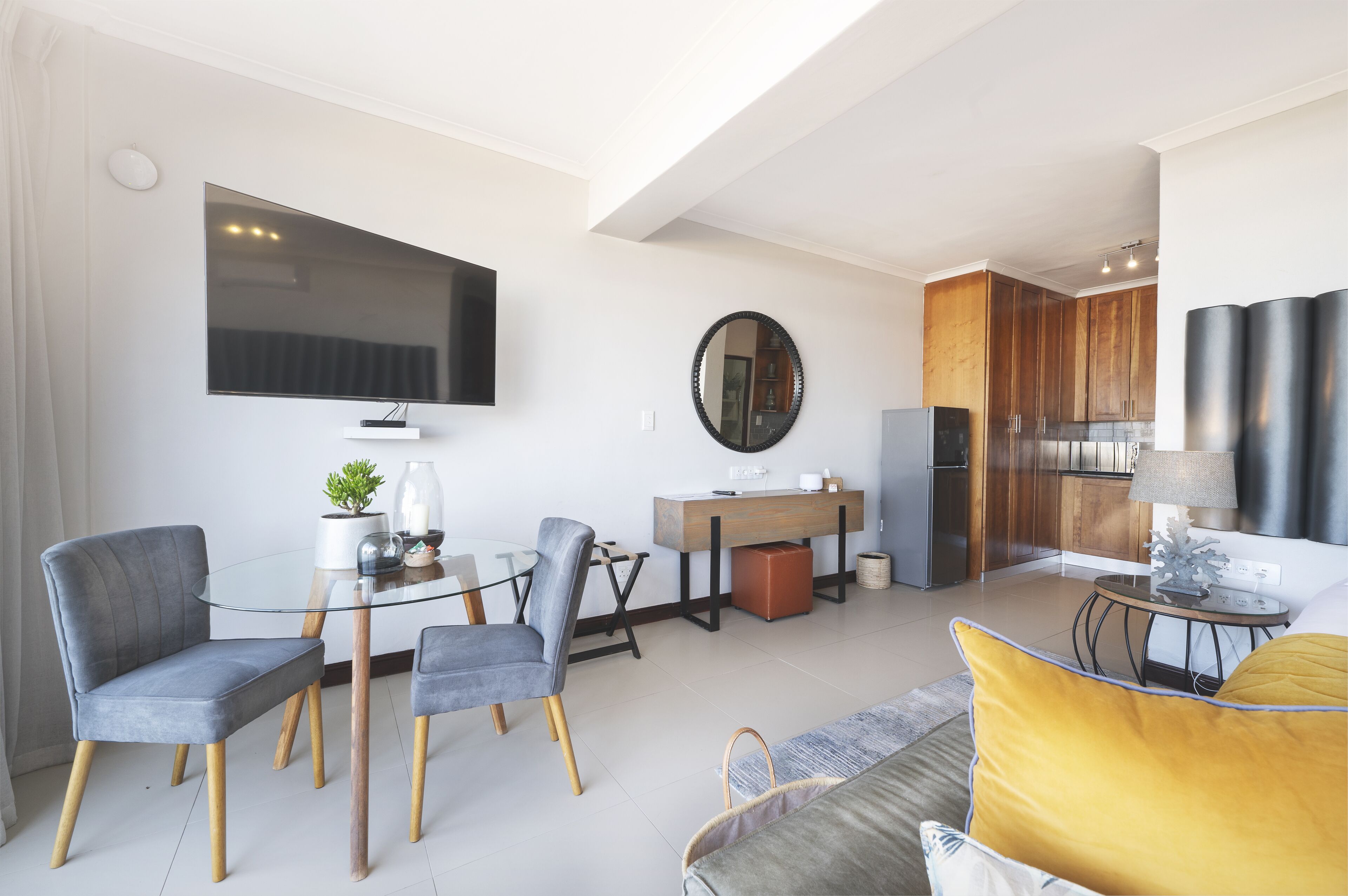 A modern living room with a flat-screen TV, a round mirror, and a dining area with a glass table and chairs.