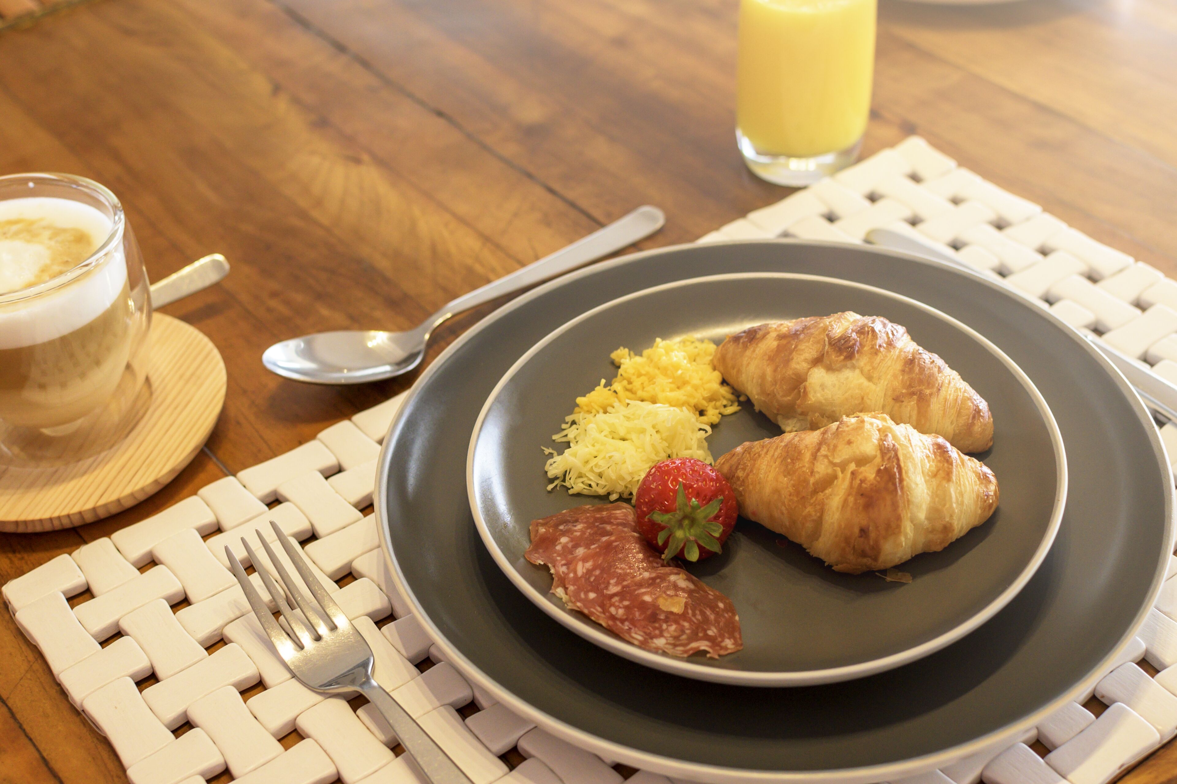 A breakfast plate with croissants, a strawberry, and scrambled eggs on a dark plate, accompanied by a glass of orange juice and a cup of coffee on a wooden table.