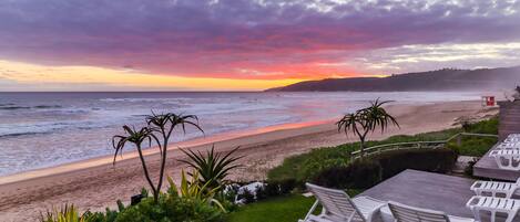 A beachside view with a sunset, a wooden deck with white lounge chairs, and a rocky shoreline.