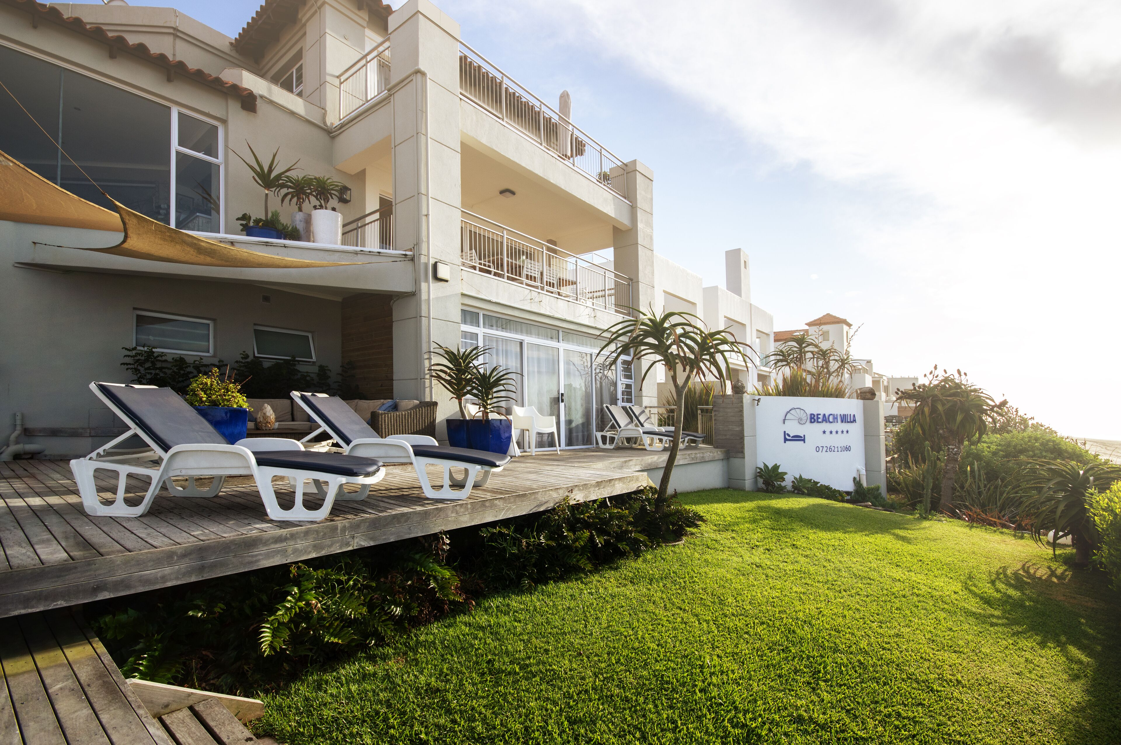 A beachfront property with a wooden deck, lounge chairs, and a sign indicating 'Beach Villa'.