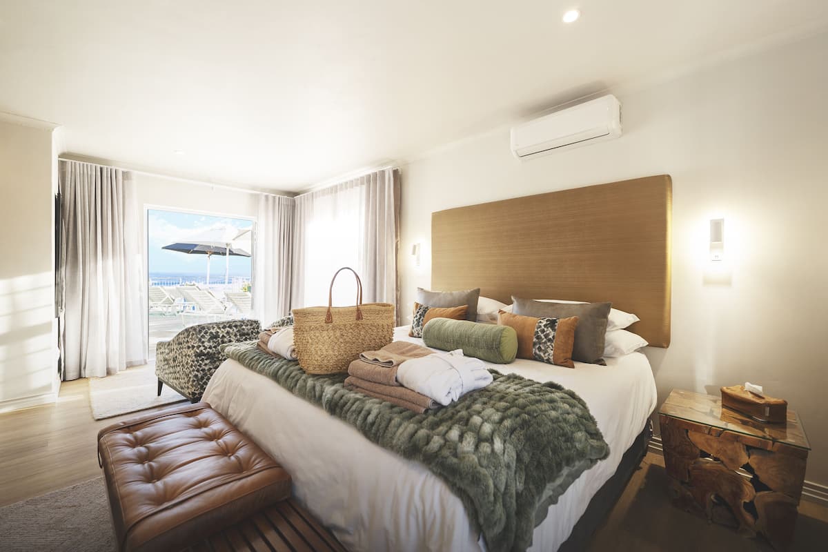 A hotel room with a large bed, a wooden headboard, a bench, a wicker basket, and a view of the beach through the window.