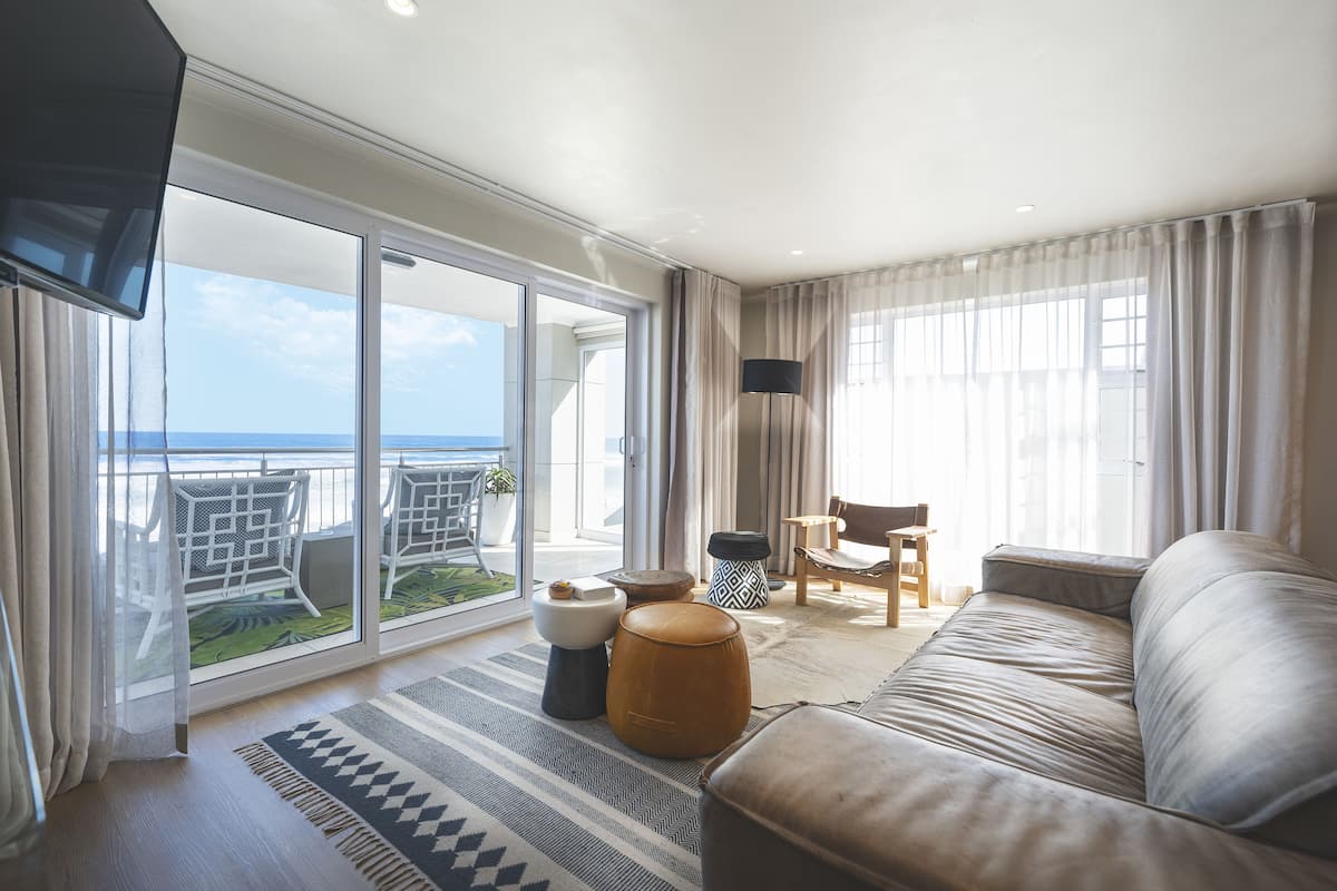 A spacious living room with a large sofa, a coffee table, and a TV mounted on the wall. There is a balcony with a view of the ocean.