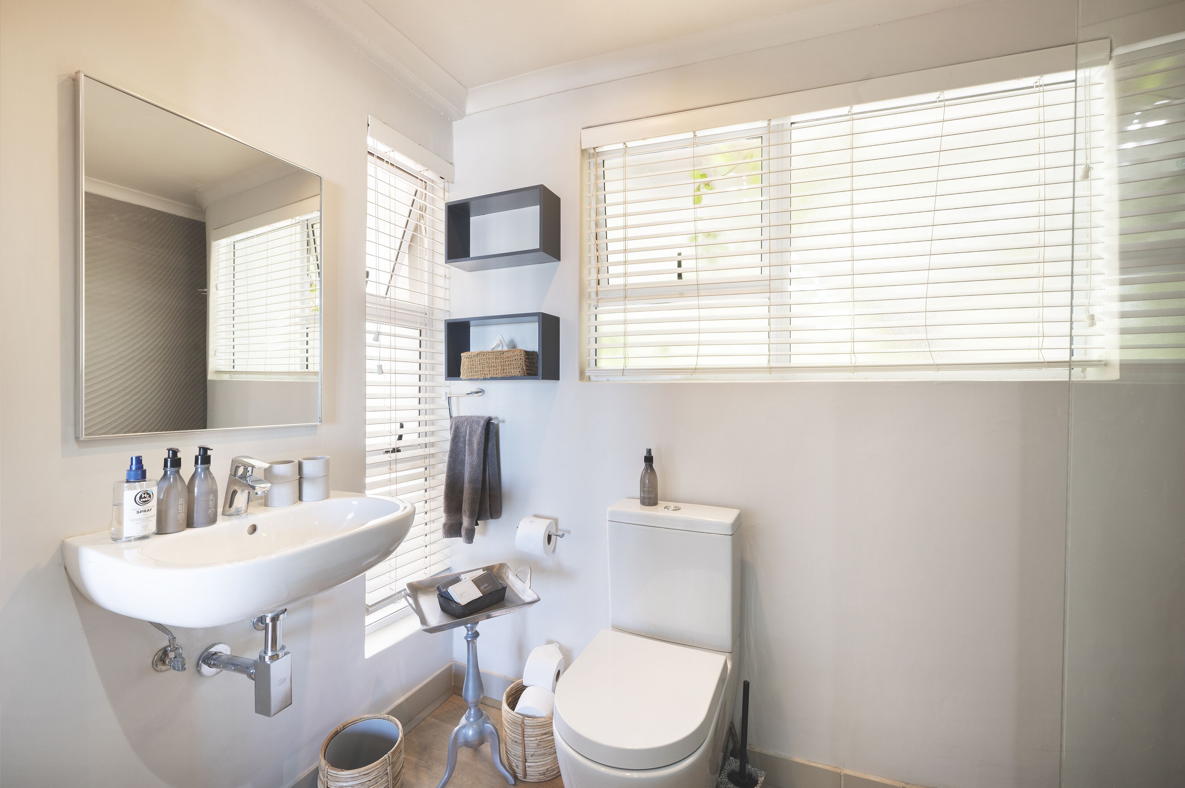 Une salle de bain moderne avec un lavabo blanc, un miroir, des toilettes et des stores aux fenêtres.