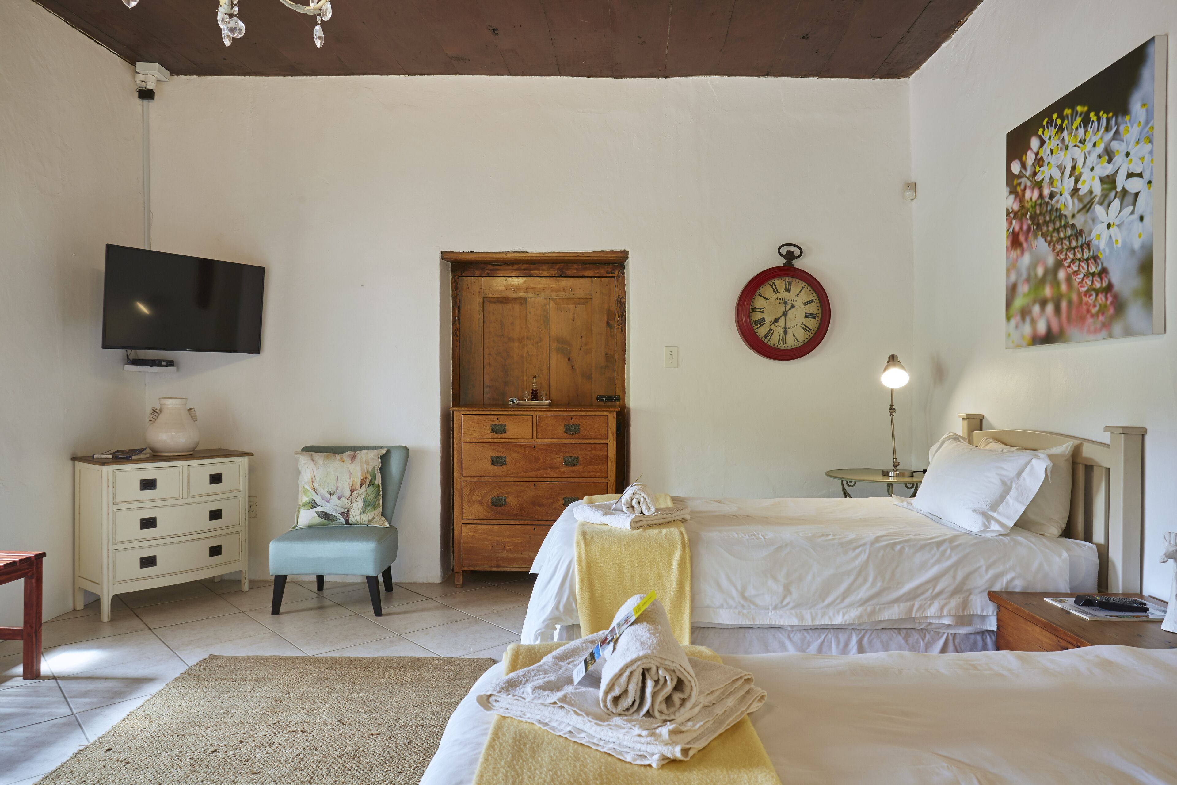 A bedroom with two beds, a wooden wardrobe, a television, a clock, and a painting.