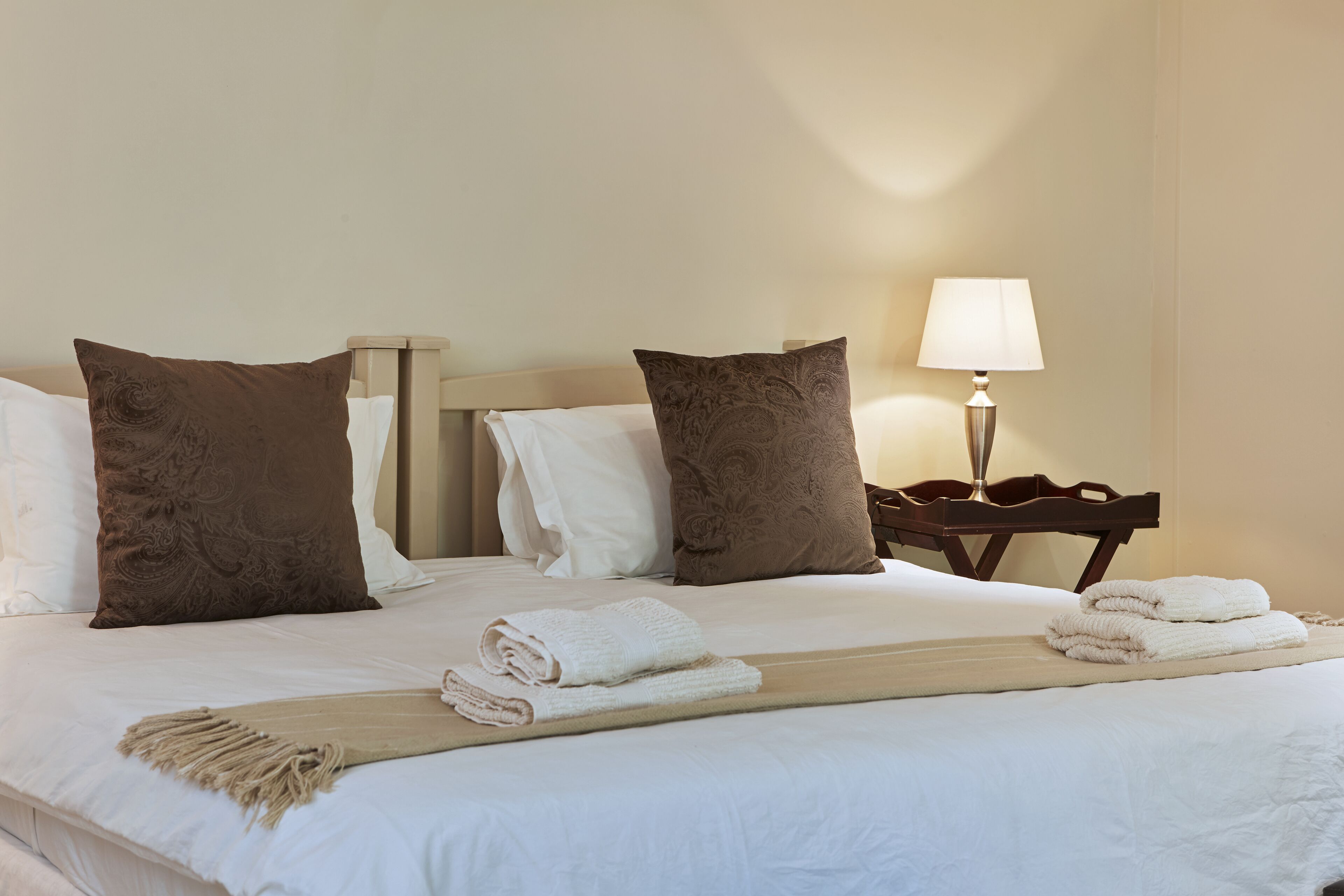 A neatly made bed with white and brown linens, a beige throw blanket, and a bedside table with a lamp and folded towels.