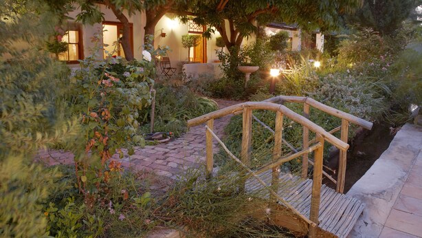 A garden with a wooden bridge, a stone pathway, and a building with multiple windows and a door. - Yellow Aloe Guesthouse (Cederberg)