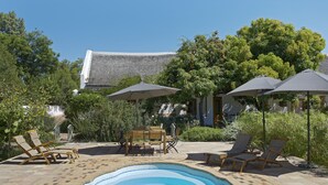 A pool area with lounge chairs and umbrellas, surrounded by greenery and a building with a thatched roof. - Yellow Aloe Guesthouse (Cederberg)