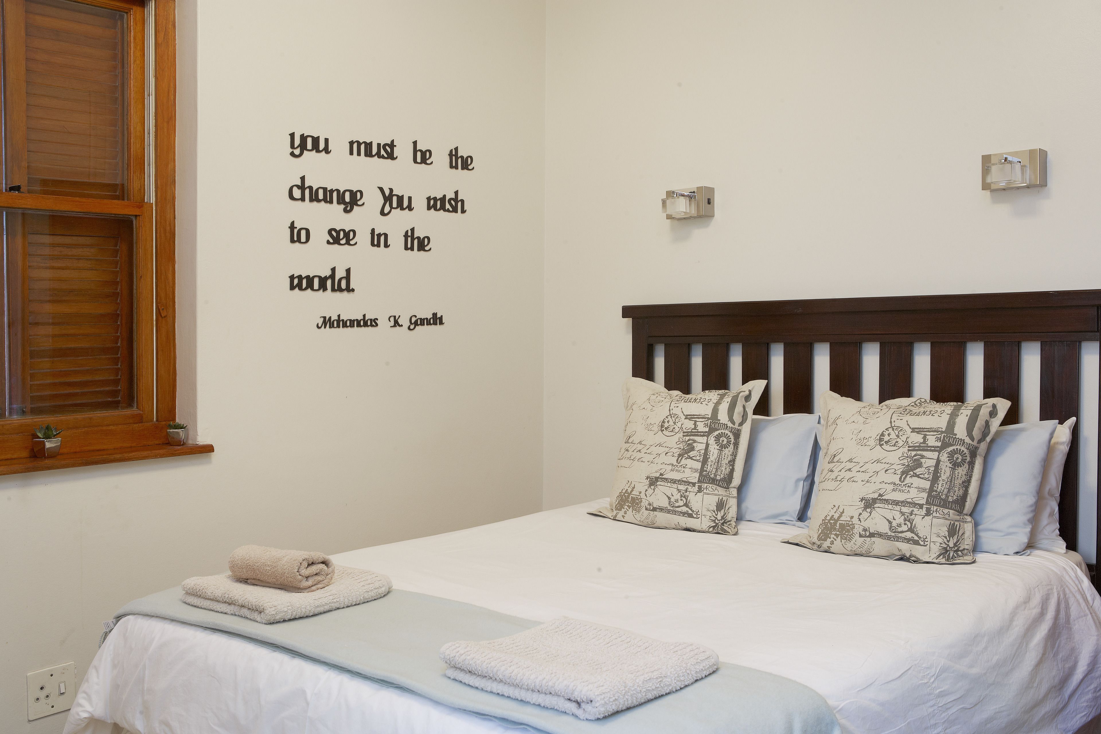 A neatly made bed with a white comforter and two pillows, a wall quote by Mahatma Gandhi, and a small window with blinds.
