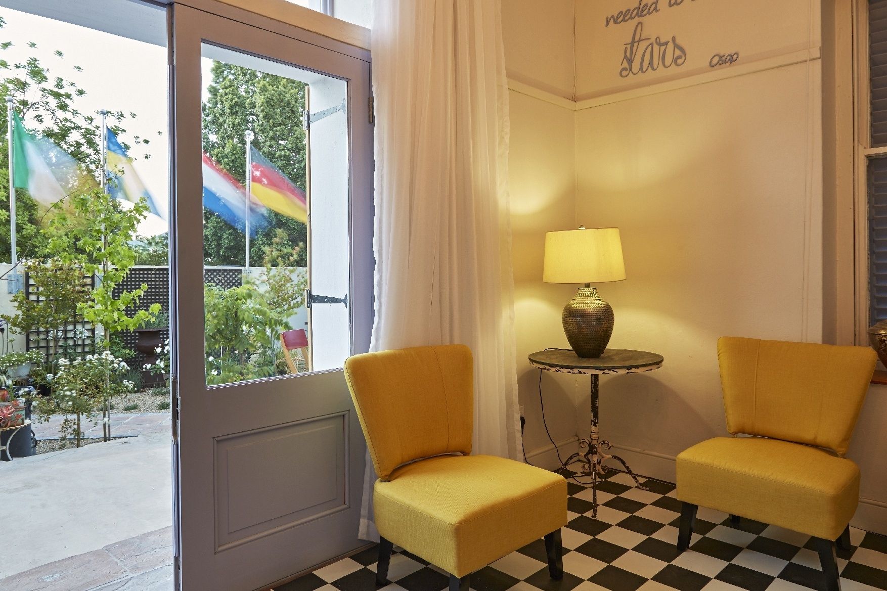 A room with two yellow chairs, a small table with a lamp, a checkered floor, and a view of a rainbow flag outside.