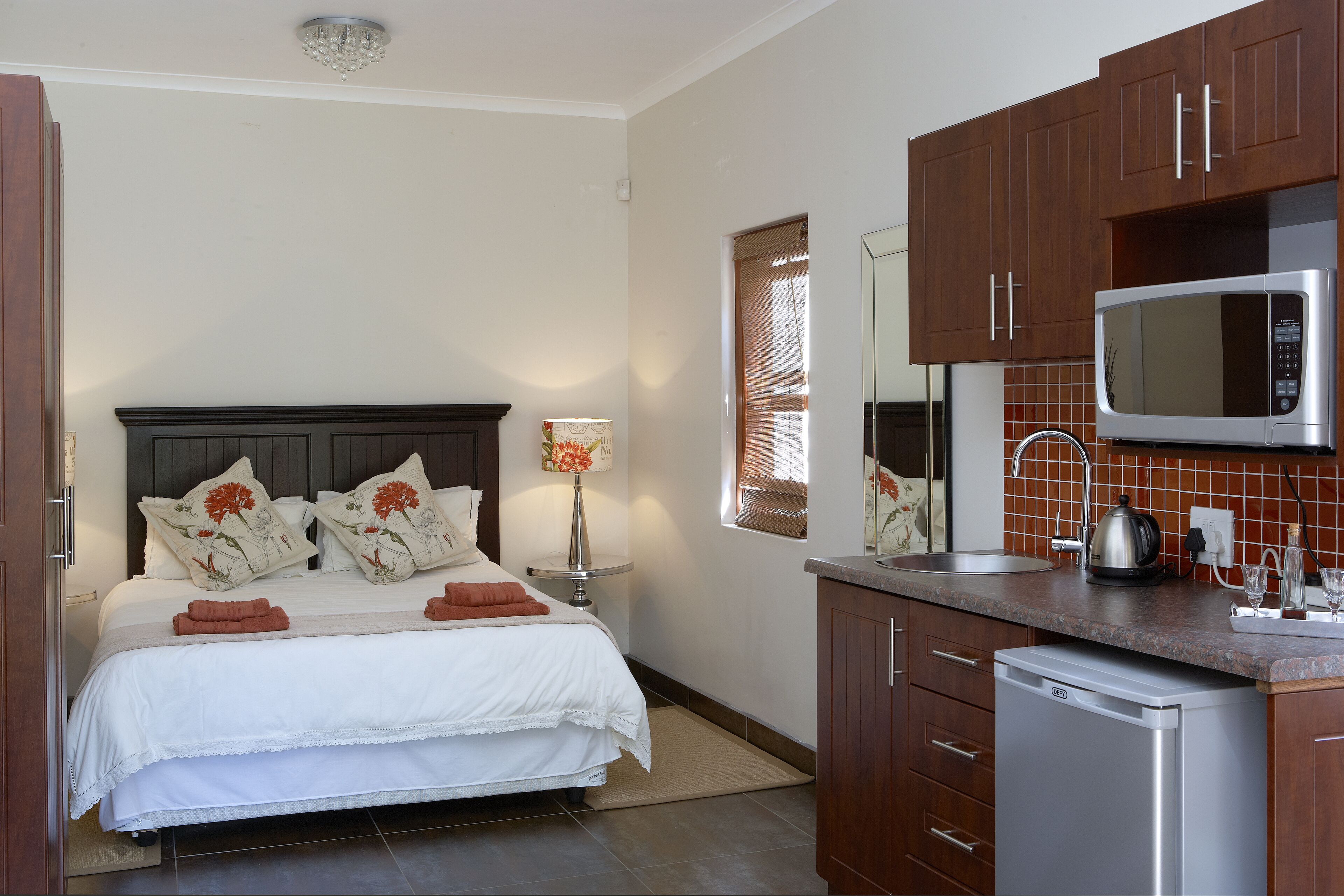 A bedroom with a bed, pillows, and a bedside lamp. Adjacent to the bedroom is a kitchen with a microwave, sink, and dishwasher.