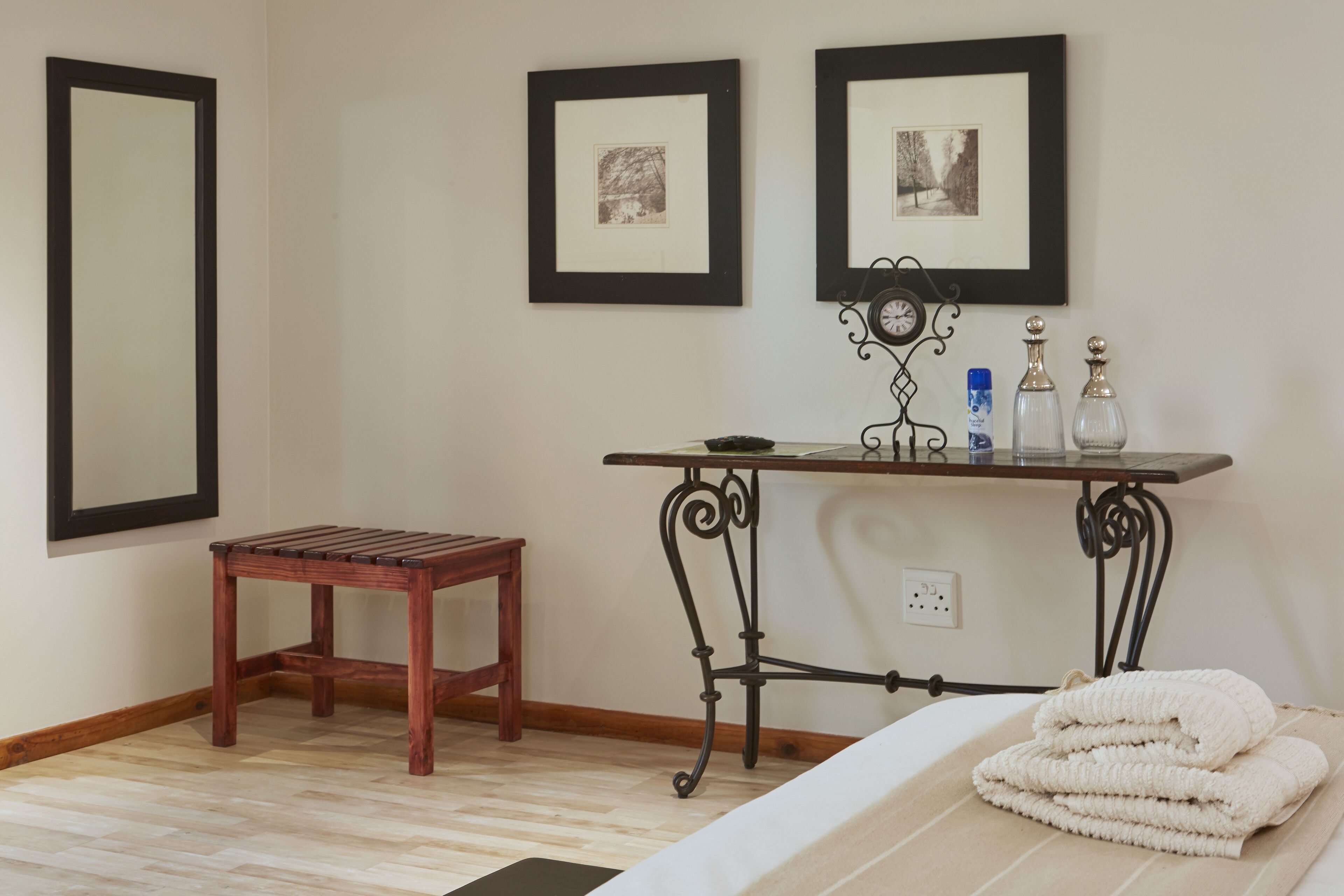 A room with a bed, a wooden stool, a table with a clock, and framed pictures on the wall.
