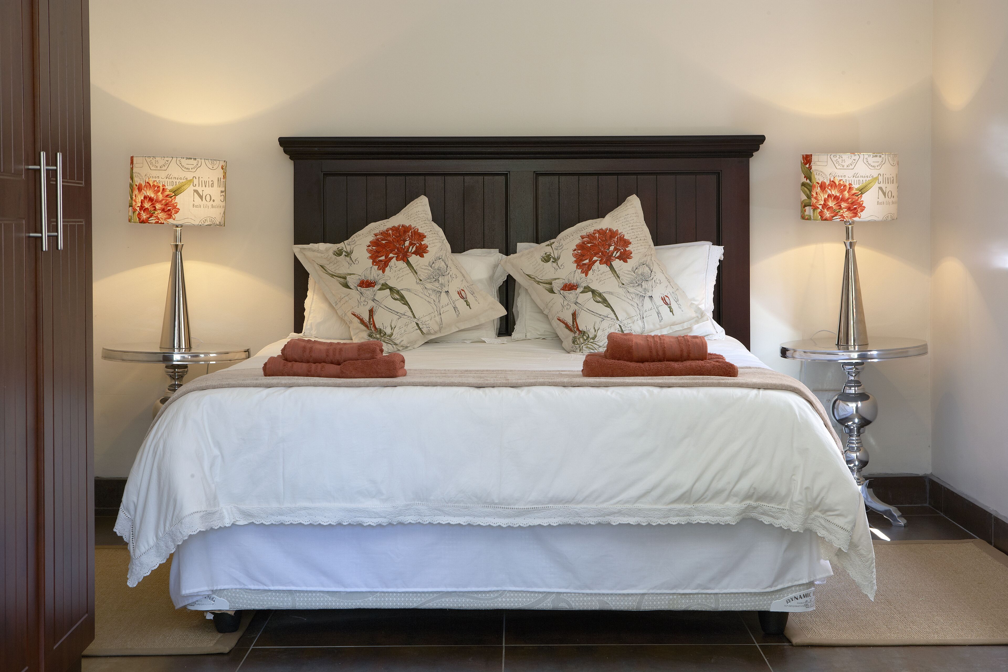 Una cama bien hecha con ropa blanca y dos almohadas decorativas, flanqueada por dos lámparas de noche con diseños florales.
