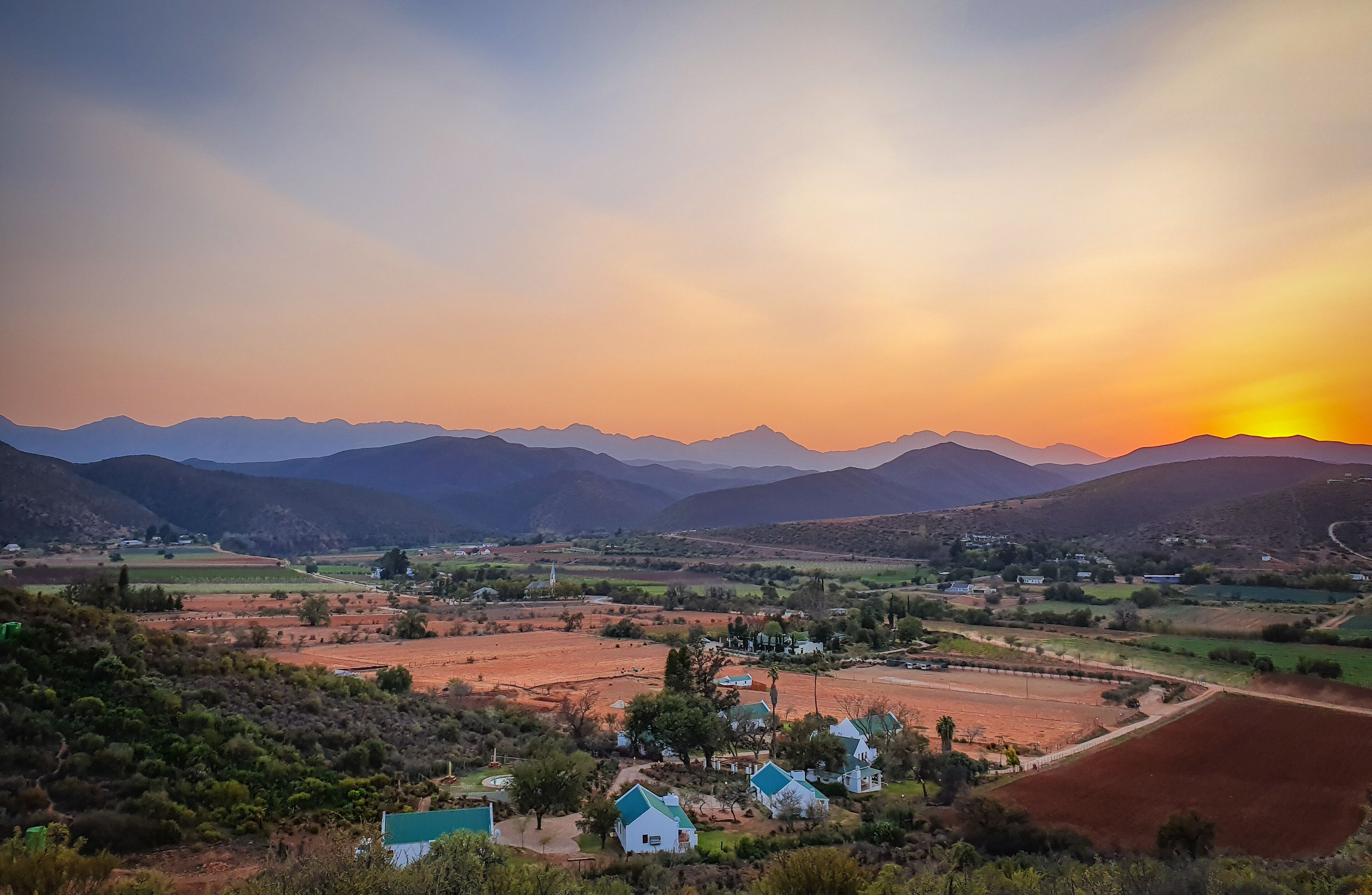 Ländliche Landschaft mit Sonnenuntergang, Bergen im Hintergrund und einem kleinen Dorf mit Häusern und Feldern.