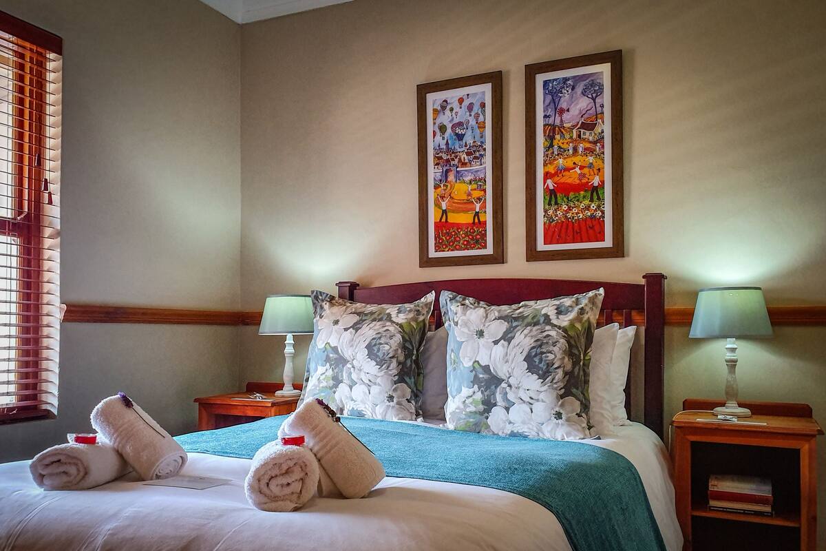 Habitación de hotel con una cama, dos almohadas, una mesita de noche con una lámpara y dos cuadros enmarcados en la pared.