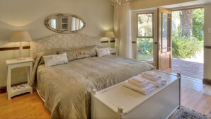 A bedroom with a large bed, a chandelier, a mirror, and a view of the garden through an open door.