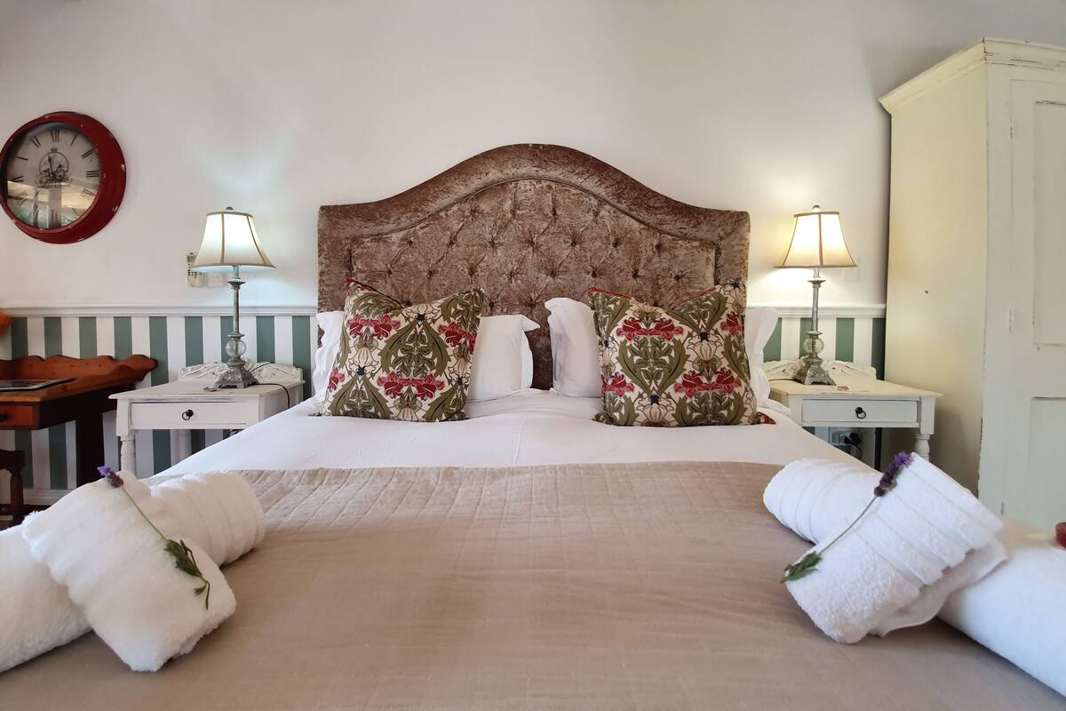 Una cama impecablemente arreglada con almohadones florales, dos lámparas de noche, un reloj y una mesita de noche de madera con lámpara.