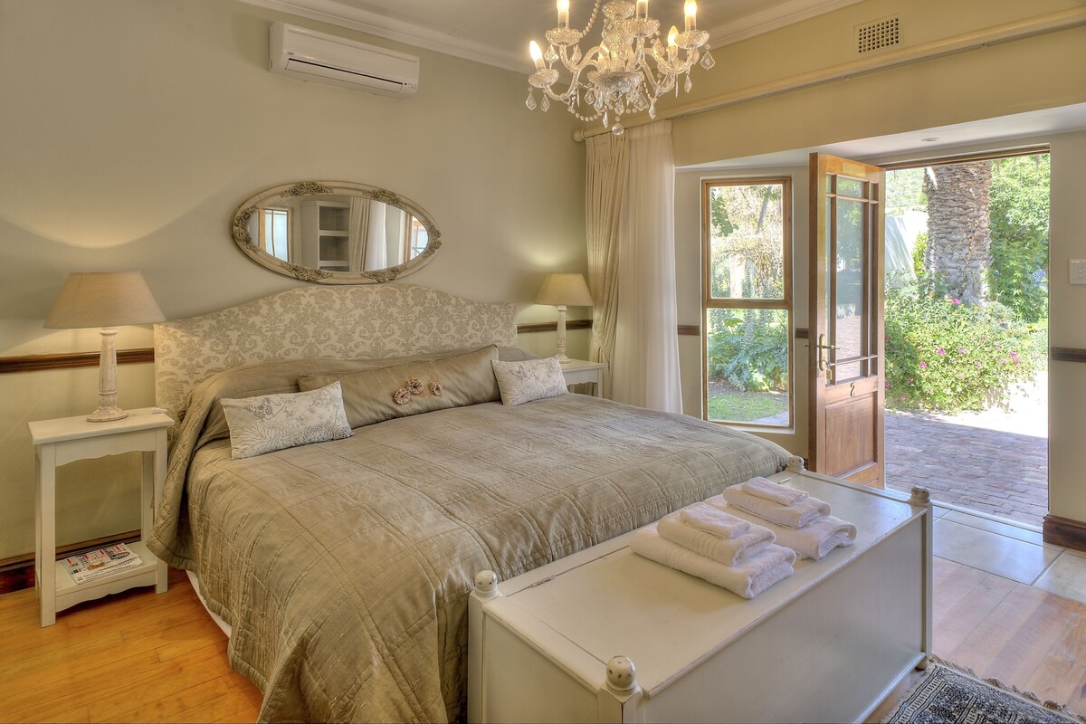 A bedroom with a large bed, a chandelier, a mirror, and a view of a garden through an open door.