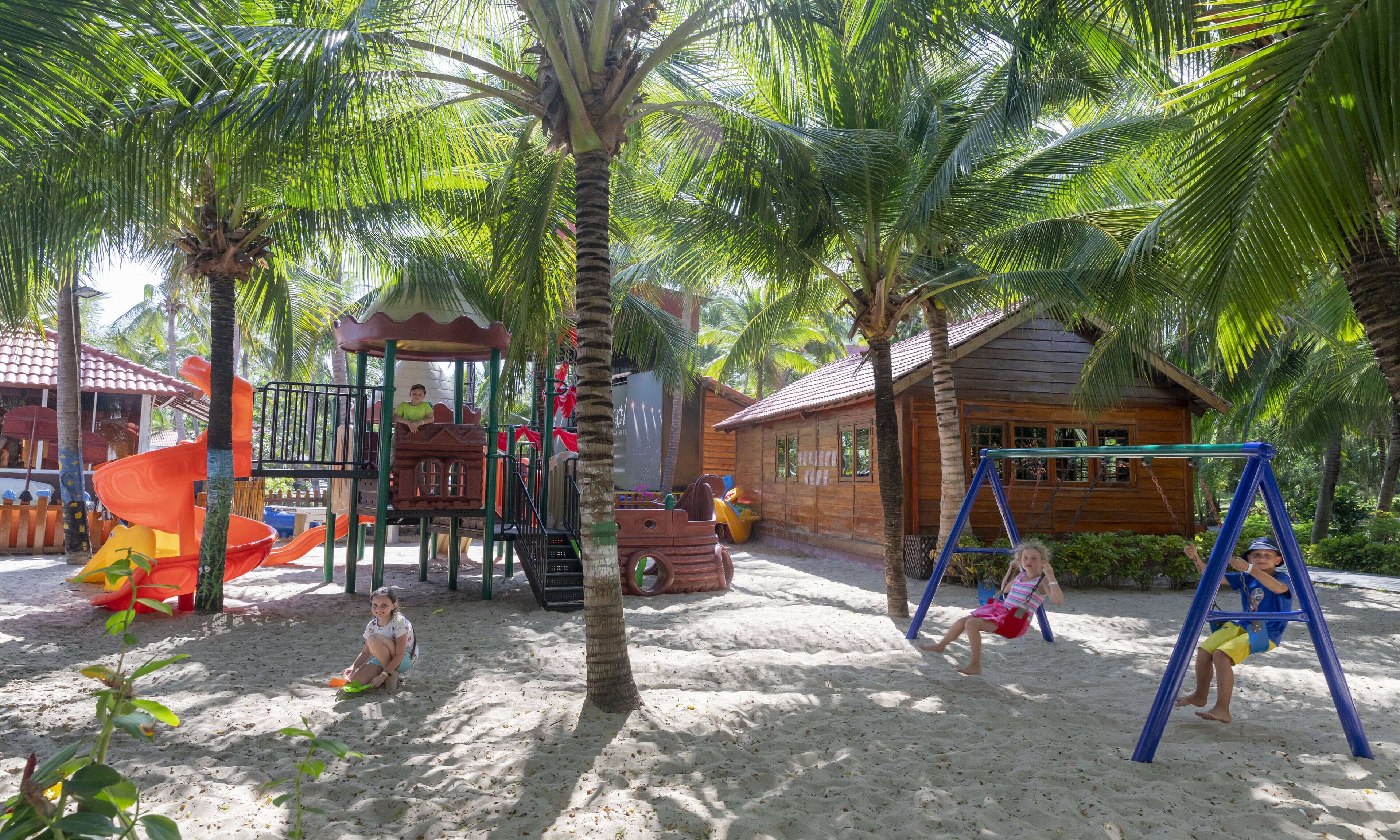 A playground with slides and swings, surrounded by palm trees and a wooden building.