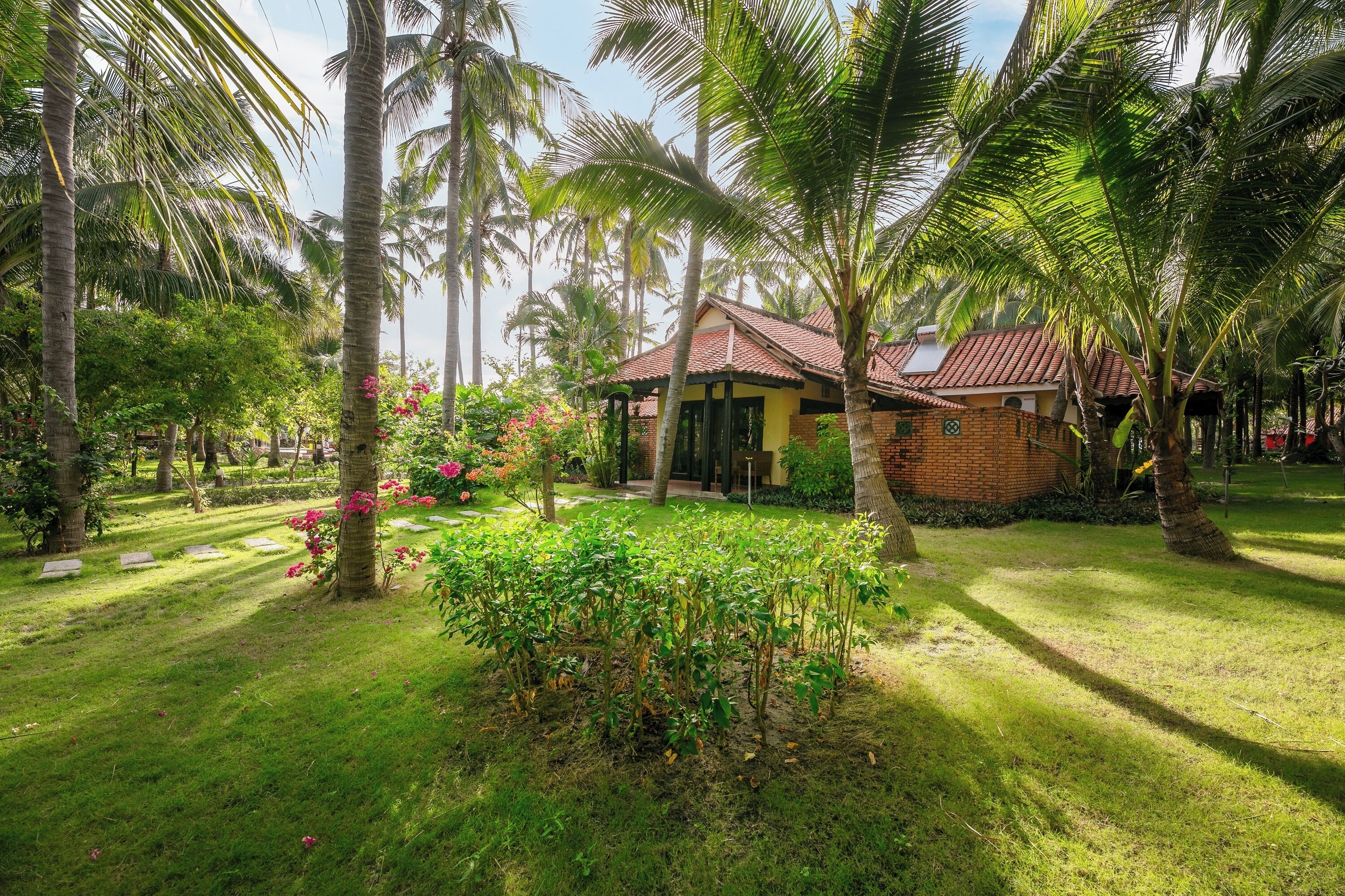 A tropical garden with palm trees, a brick house, and a well-maintained lawn.
