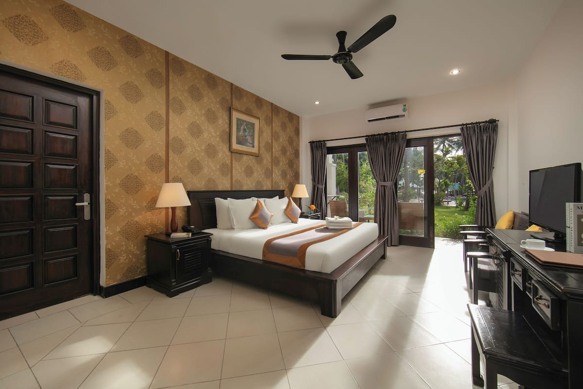 Una habitación de hotel con una cama grande, dos mesitas de noche con lámparas, un escritorio con una silla, un ventilador de techo y un ventanal con cortinas.