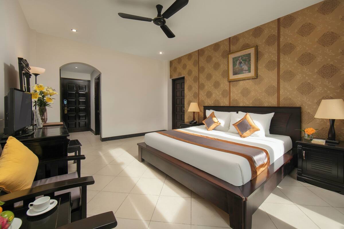 Habitación de hotel con una cama grande, un escritorio con una silla, un ventilador de techo y una puerta que da a otra habitación.