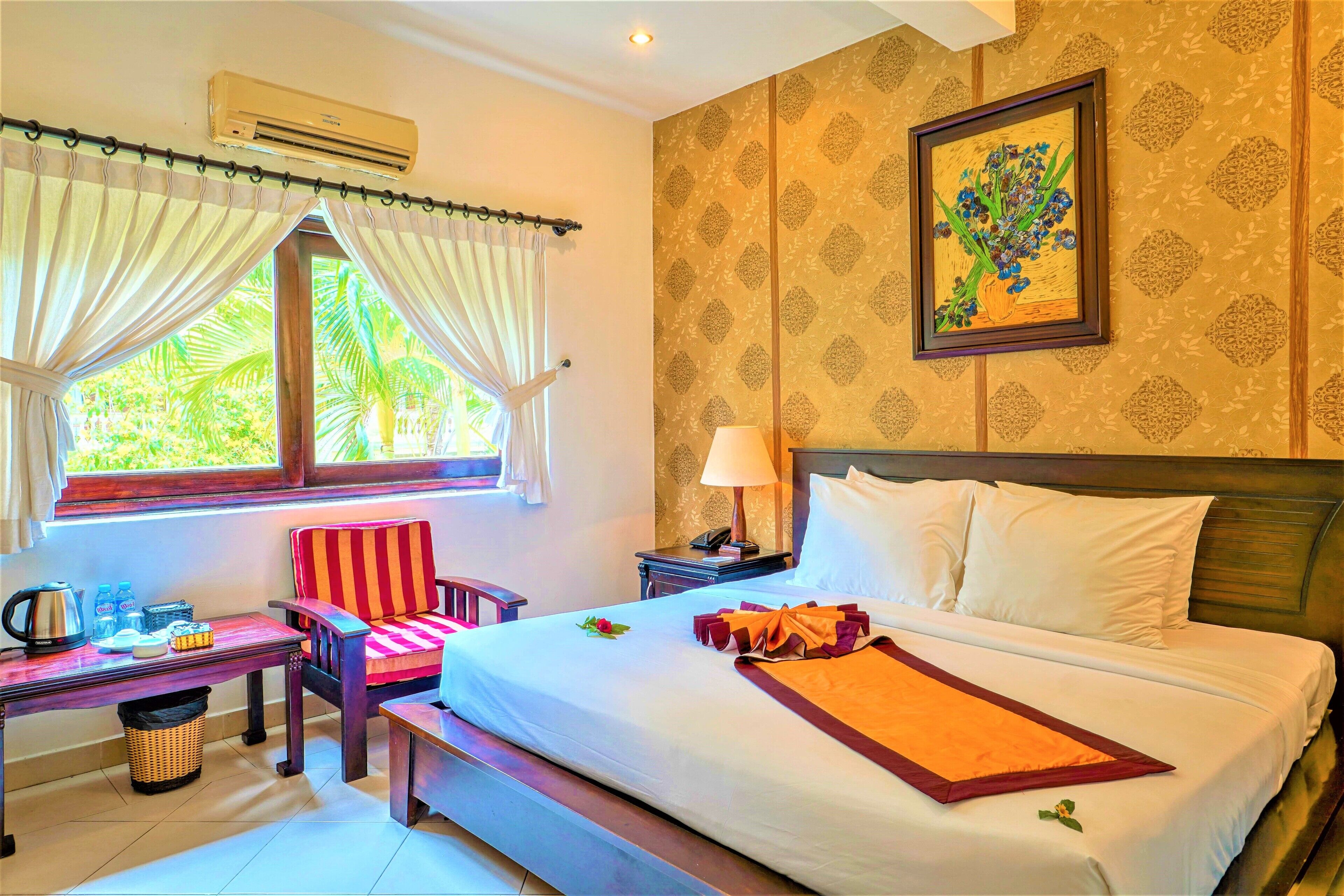 A hotel room with a bed, a chair, a table with amenities, a window with curtains, and a wall with a framed picture.