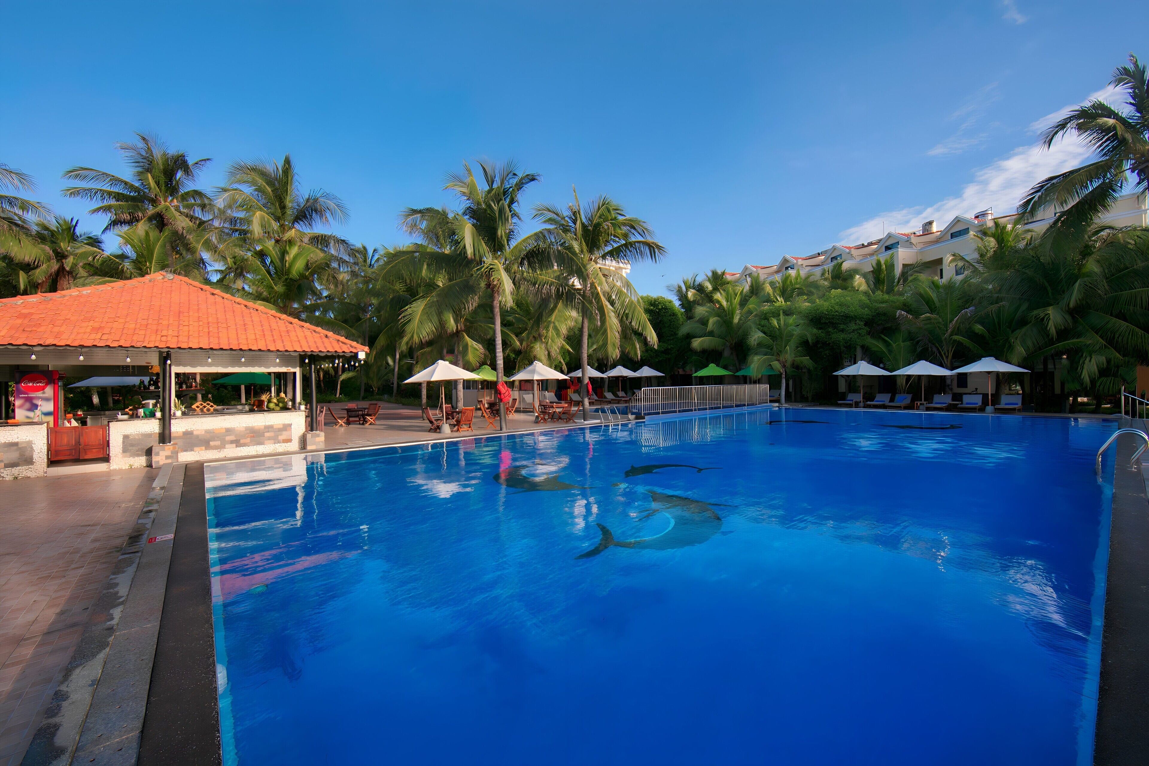 A swimming pool with palm trees and a covered area with umbrellas.