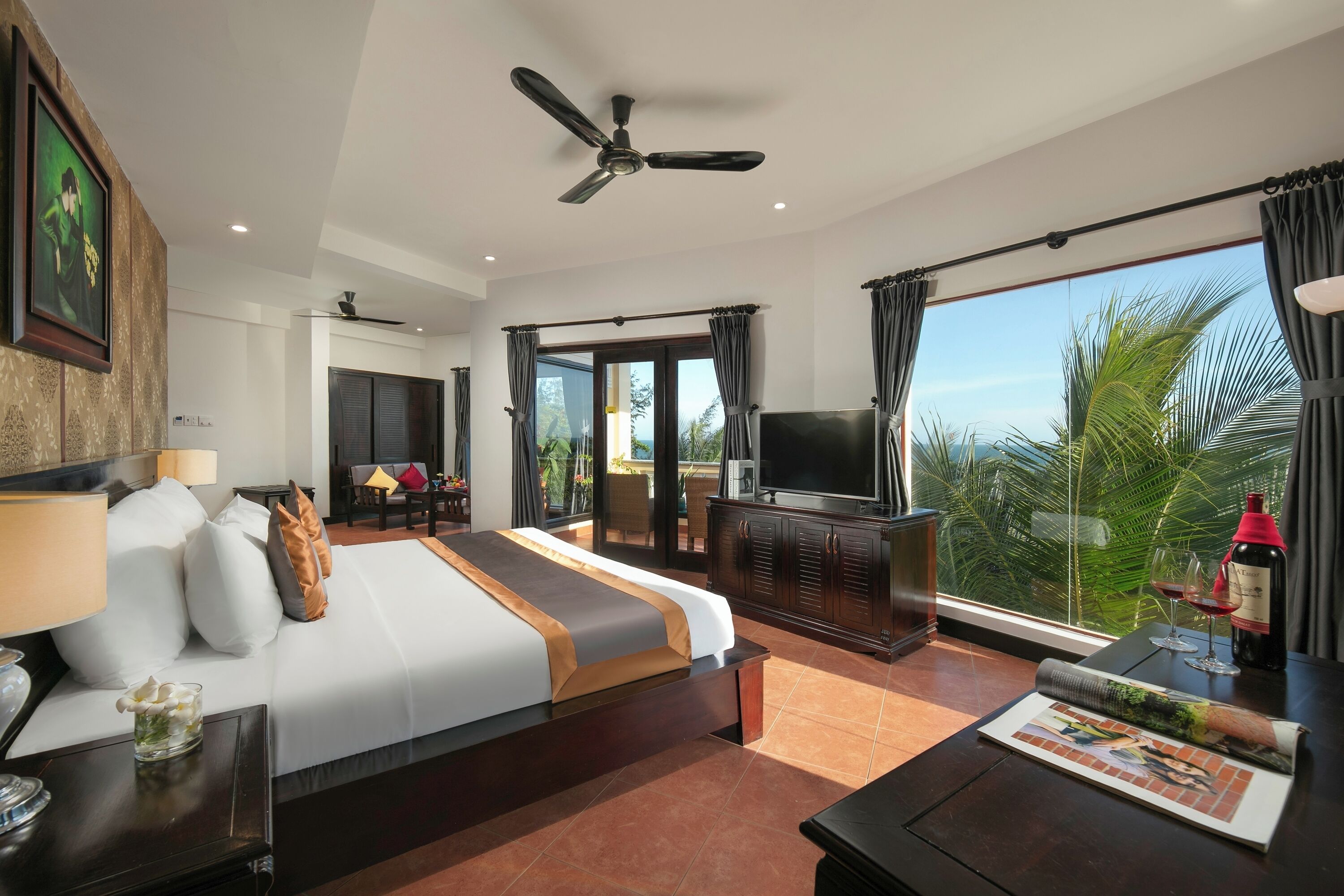A hotel room with a large bed, a TV, a balcony with a view, and a ceiling fan.