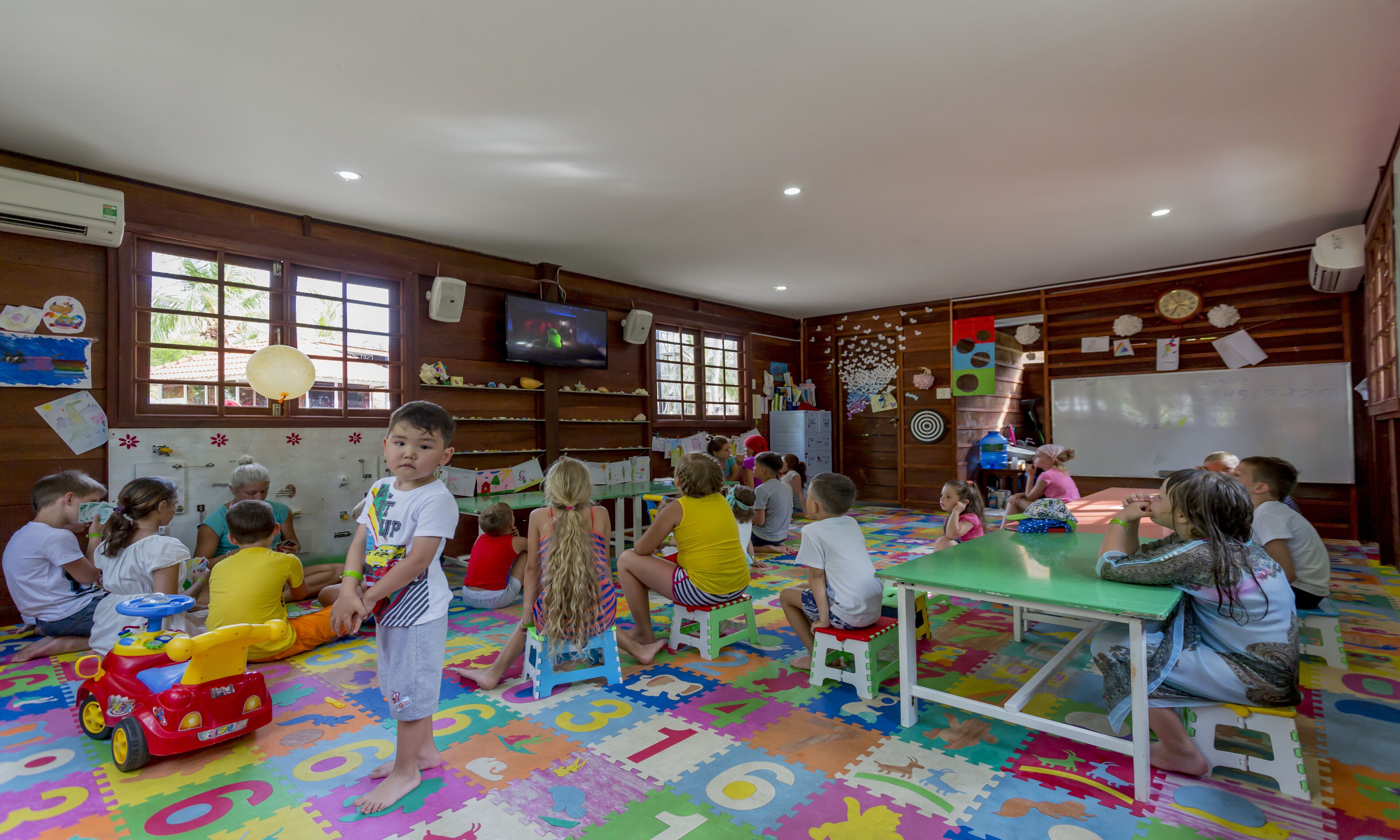 A classroom with children engaged in activities, wooden walls, a whiteboard, and educational posters.