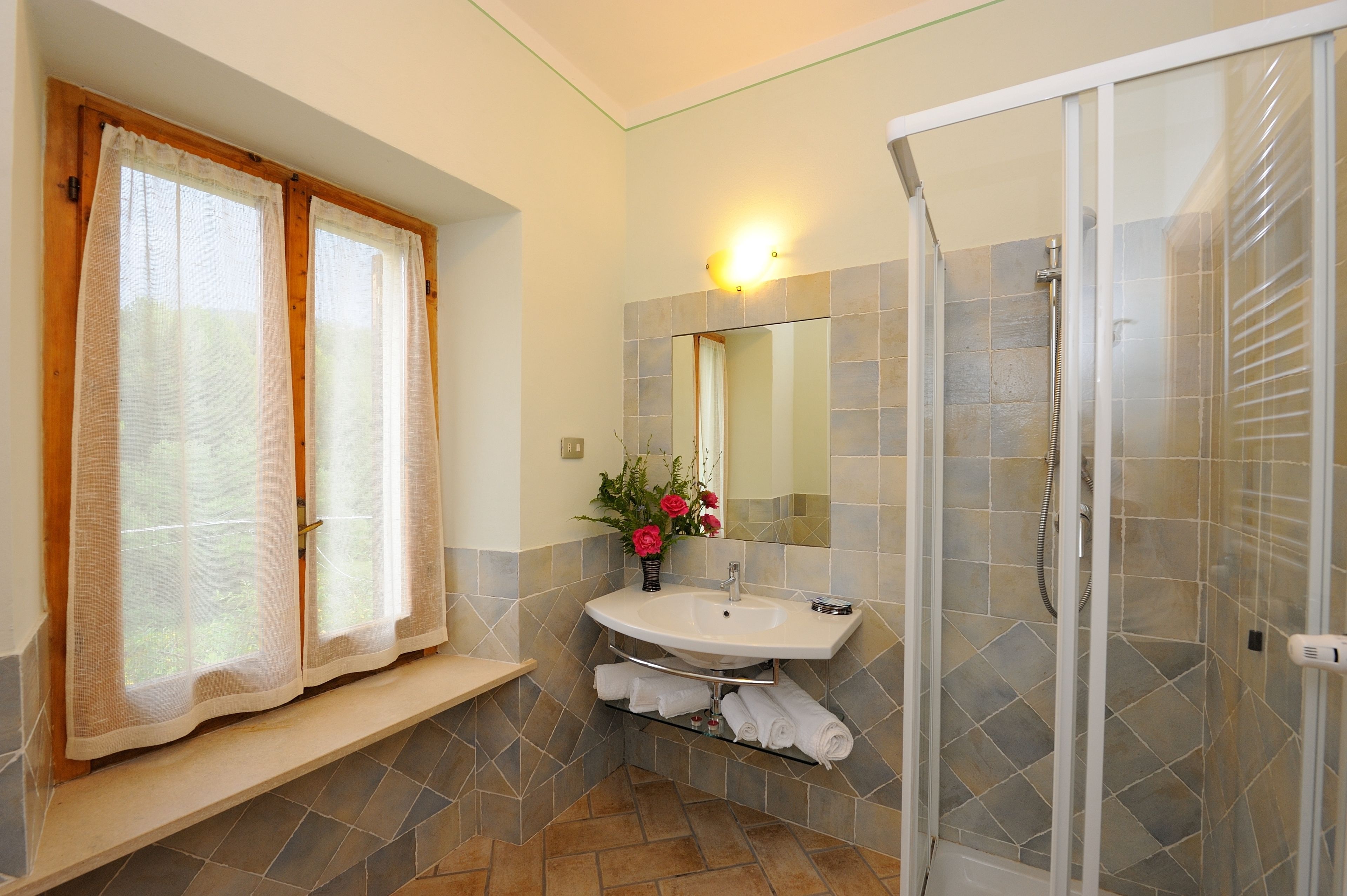 Een badkamer met een wastafel, spiegel, douche en raam met gordijnen.