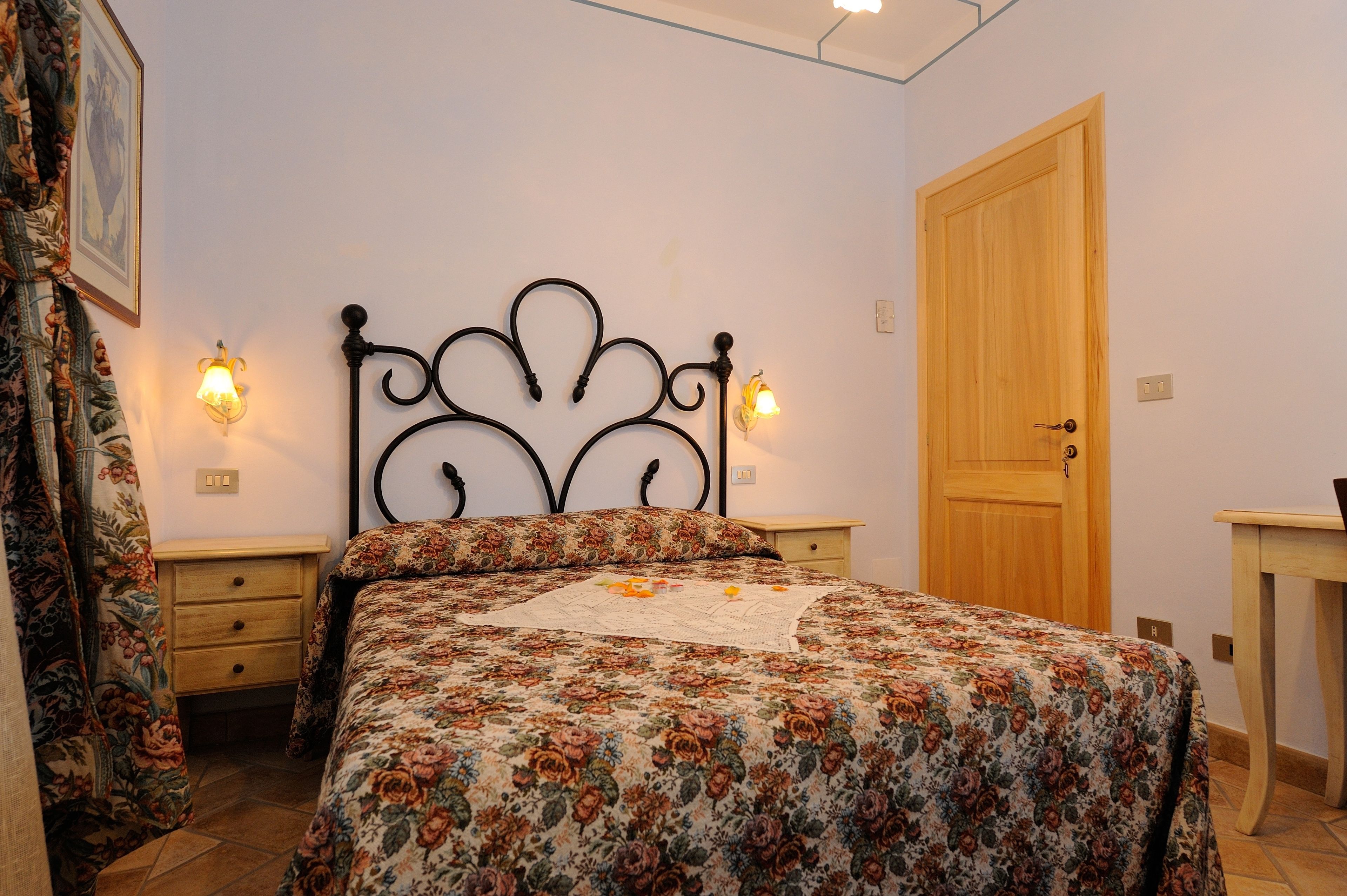 Un dormitorio con una colcha estampada floral, dos mesitas de noche con lámparas, una puerta de madera y una pequeña mesita auxiliar de madera.