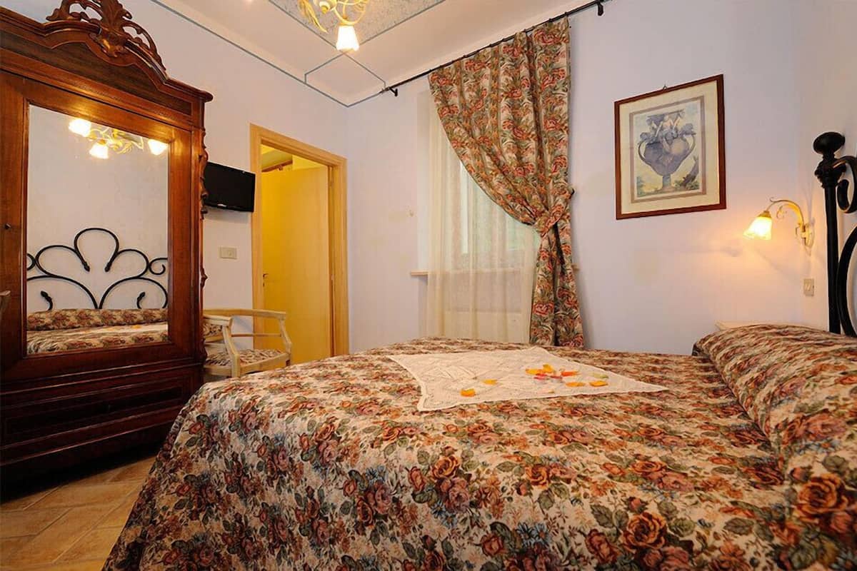 Une chambre à coucher avec une couvre-lit à motifs floraux, une armoire en bois, une chaise et un tableau accroché au mur.
