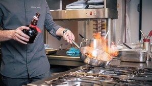 A chef in a kitchen with a stove, pots, and a bottle of sauce. - Hotel Goldene Rose (Monguelfo)
