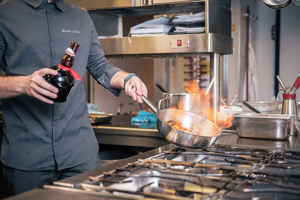 A chef in a kitchen with a stove, pots, and a bottle of sauce. - Hotel Goldene Rose (Monguelfo)