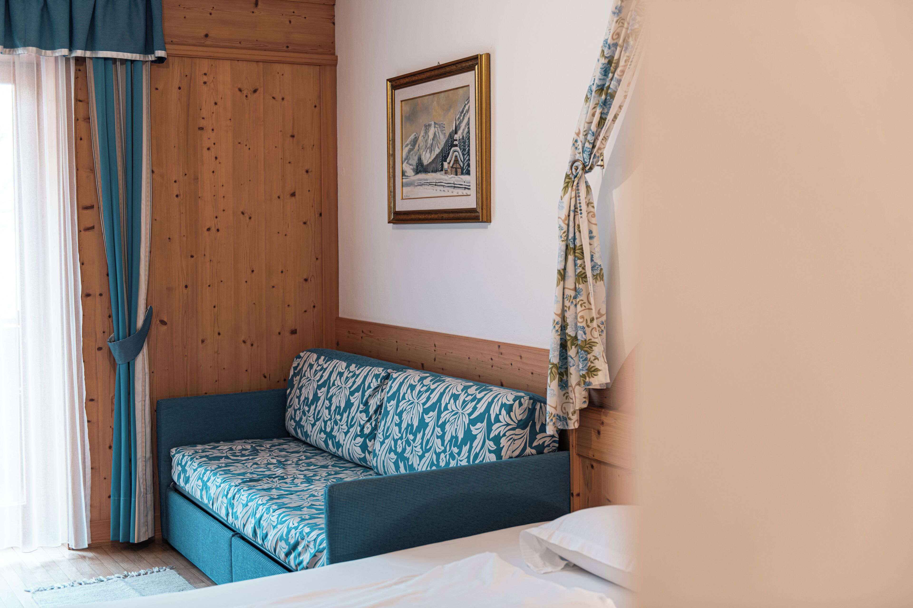 Ein Zimmer mit einer holzverkleideten Wand, einem Bett mit weißer Bettwäsche, einem blau gemusterten Sofa und einem gerahmten Bild einer Berglandschaft.