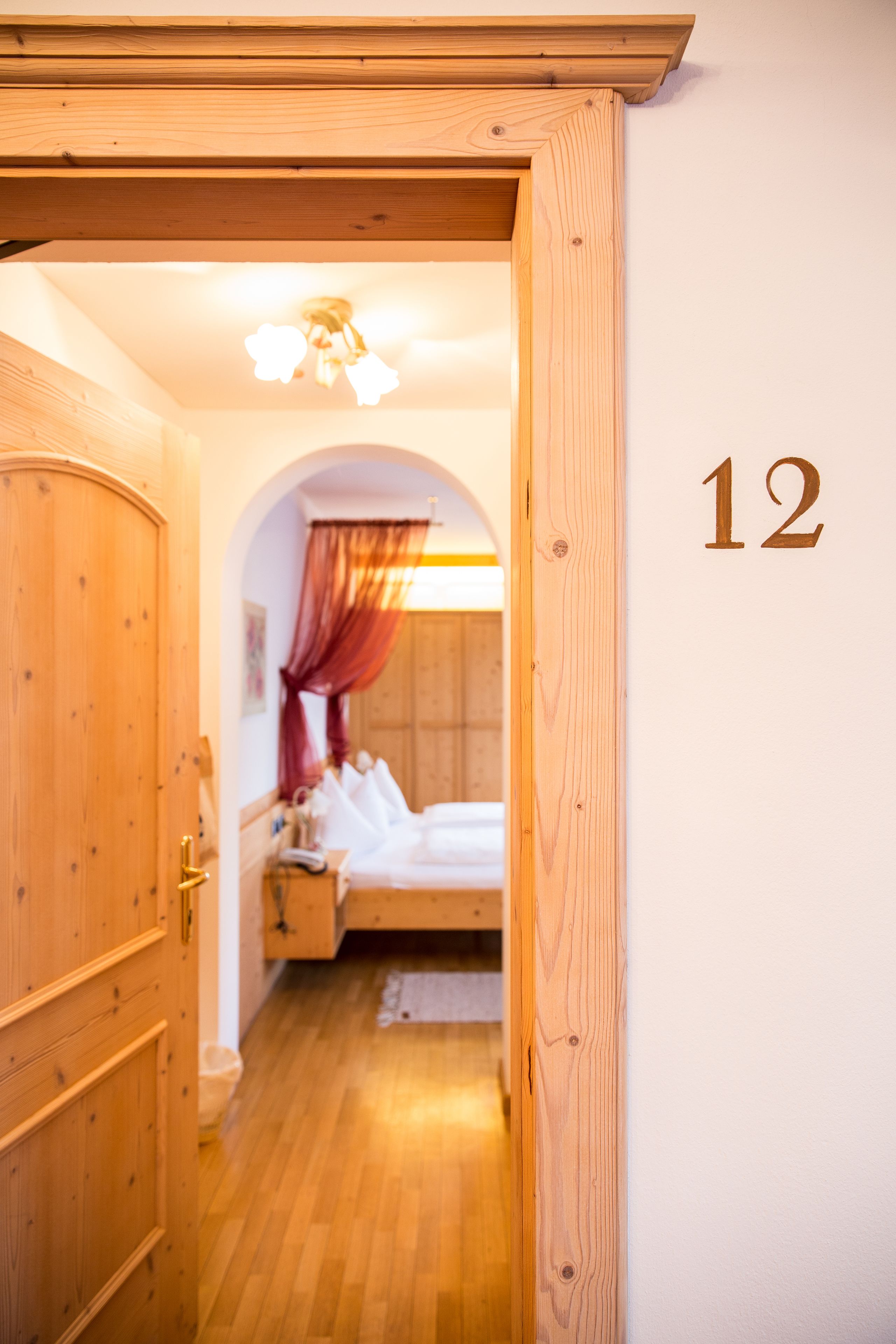 Ein Hotelzimmer mit Holztür, der Wand ist die Nummer 12 angebracht, ein Bett mit weißer Bettwäsche und ein Nachttisch mit Lampe.