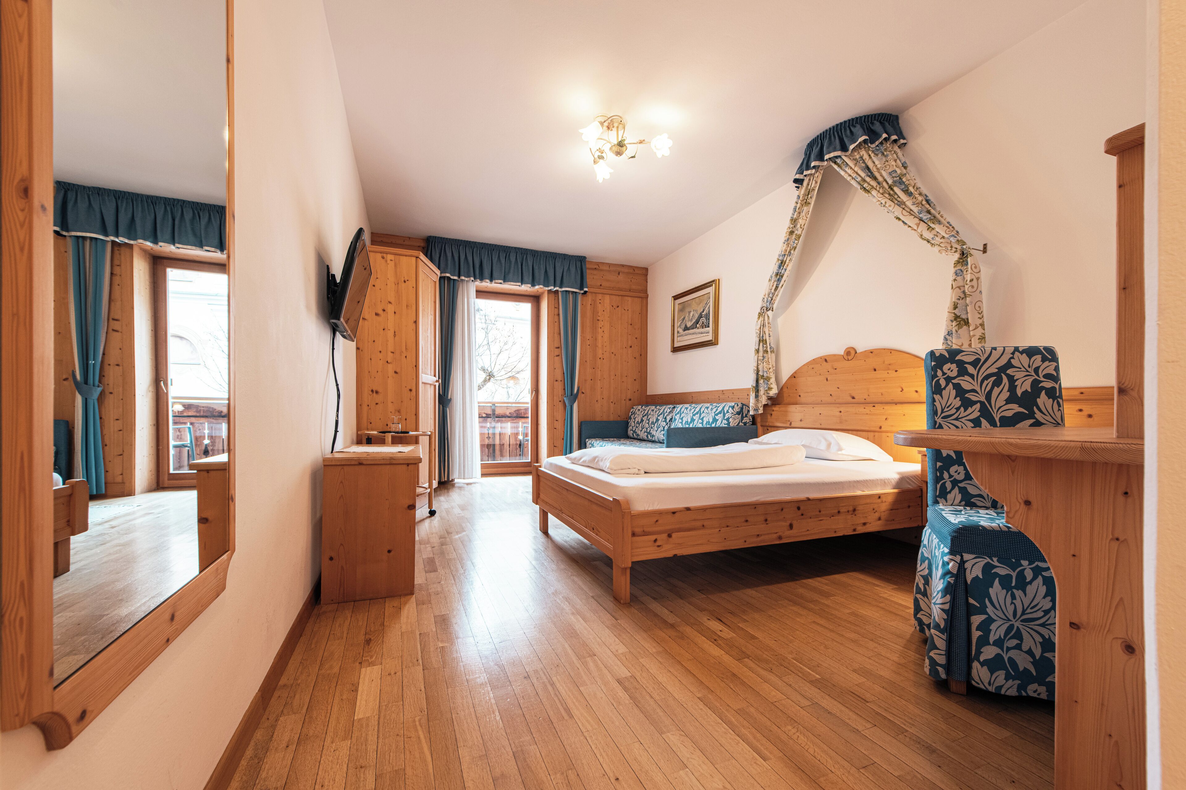 Quarto com piso de madeira, uma cama, uma cadeira, uma escrivaninha e janela com cortinas.