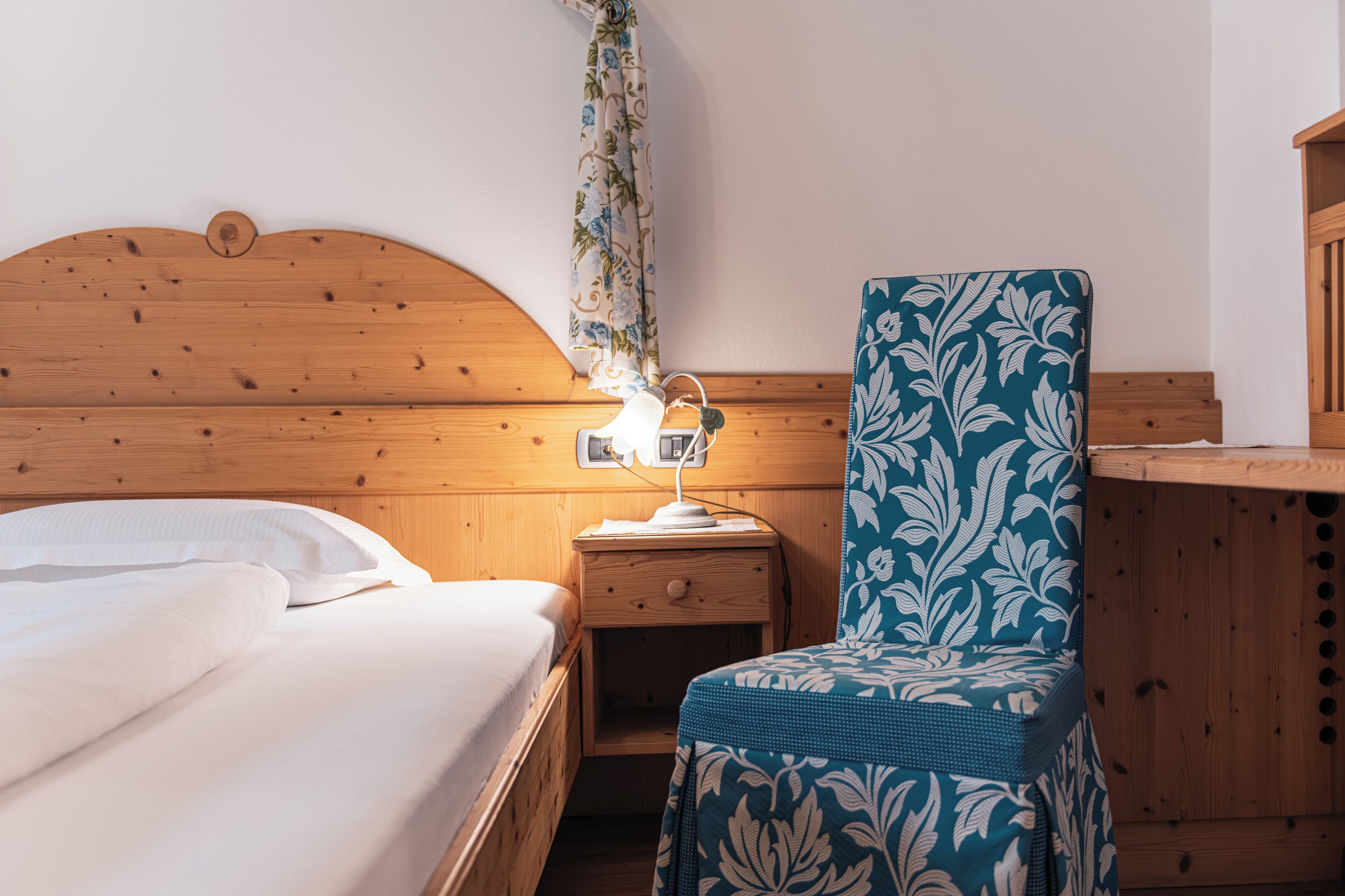 Una cama de madera con sábanas blancas, una mesita de noche con una lámpara y una silla estampada.
