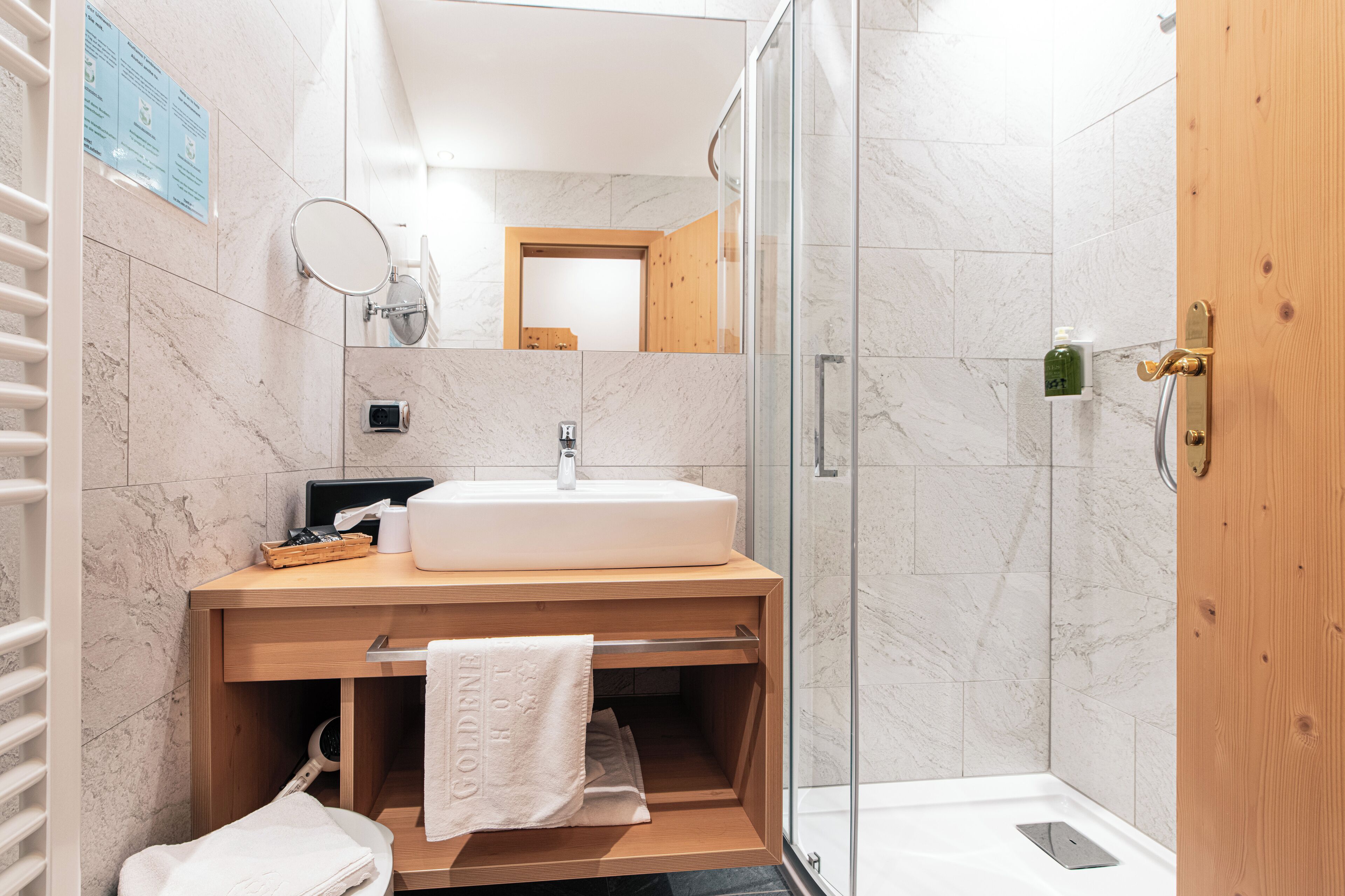 A modern bathroom with a marble-tiled wall, a wooden vanity with a sink, a glass shower enclosure, and a wooden door.