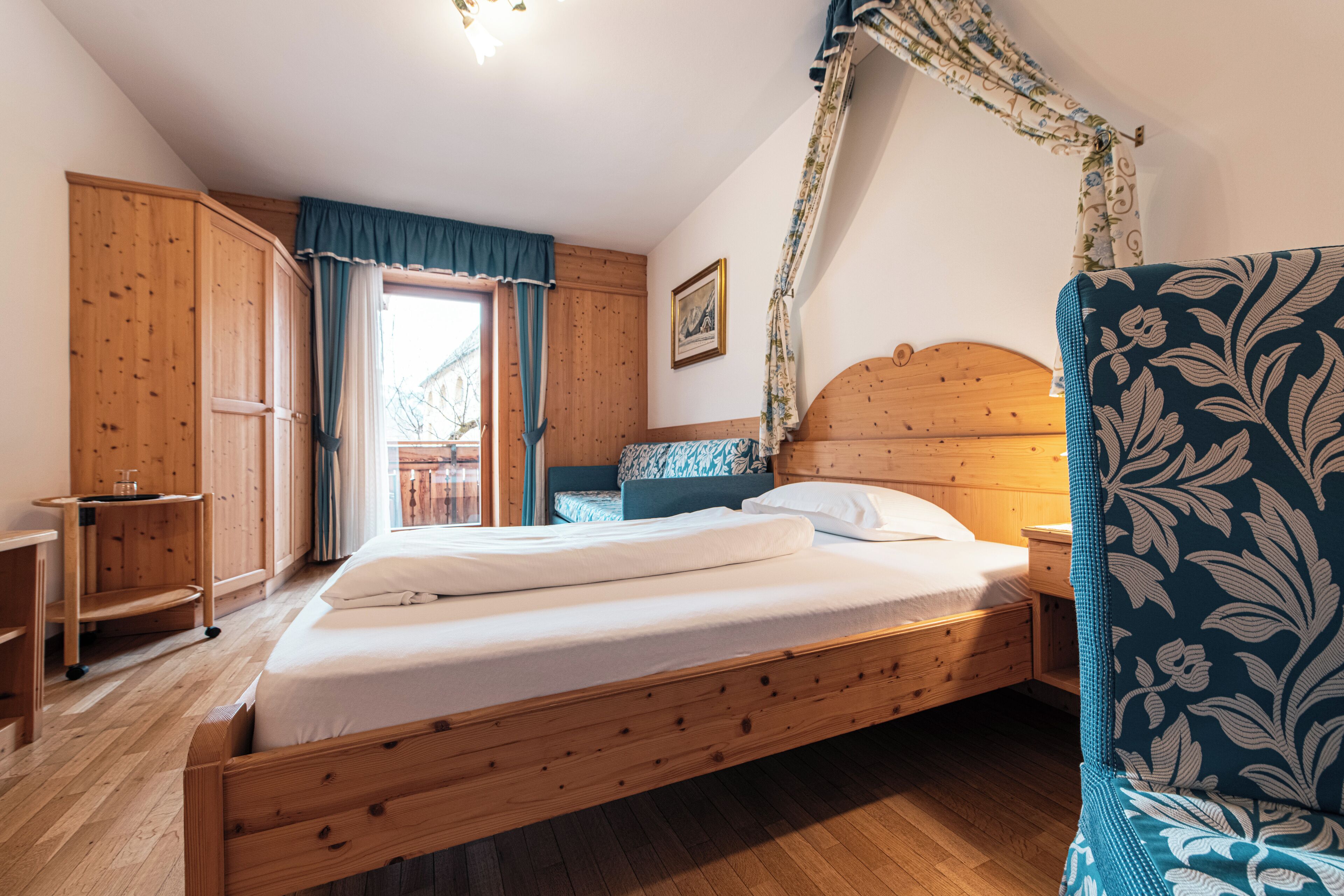 Uma cama de madeira com dossel, um guarda-roupa de madeira, uma mesinha e uma poltrona estampada num quarto com soalho e paredes de madeira.