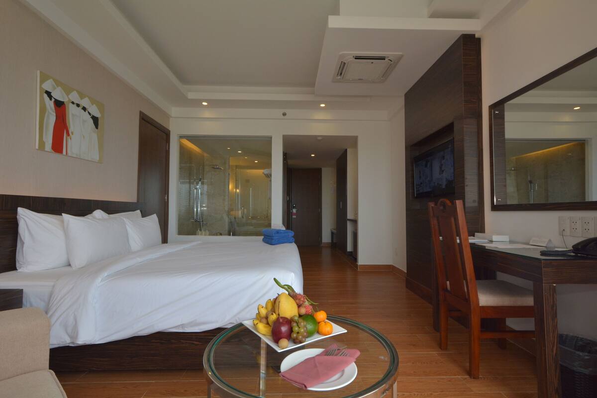 A hotel room with a bed, a glass table with fruit and a plate, a desk with a chair, and a TV.