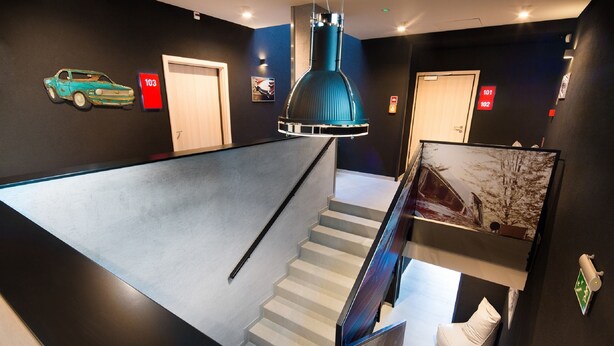 A modern hotel lobby with a staircase, a teal car artwork, and a large pendant light. - Automobil Hotel (Kraków)
