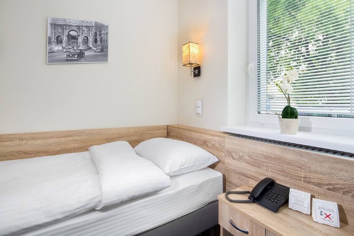 Una habitación de hotel con una cama, teléfono en la mesita de noche, luz de pared, ventana con persianas y una pequeña planta en maceta.
