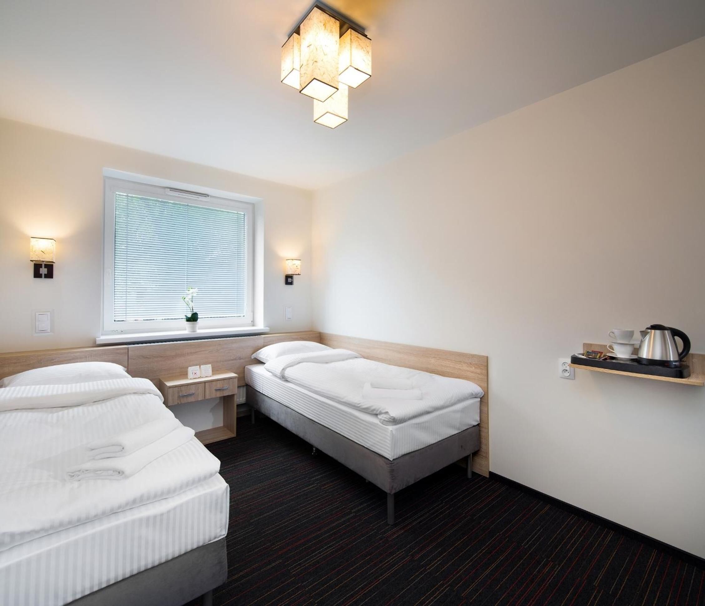 Habitación de hotel con dos camas, una ventana con persianas, una mesita de noche con lámpara y un estante con hervidor.