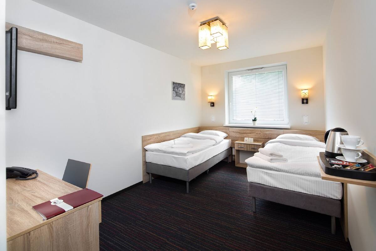 Habitación de hotel con escritorio, dos camas individuales, una mesita con una bandeja de artículos de desayuno y una ventana con persianas.