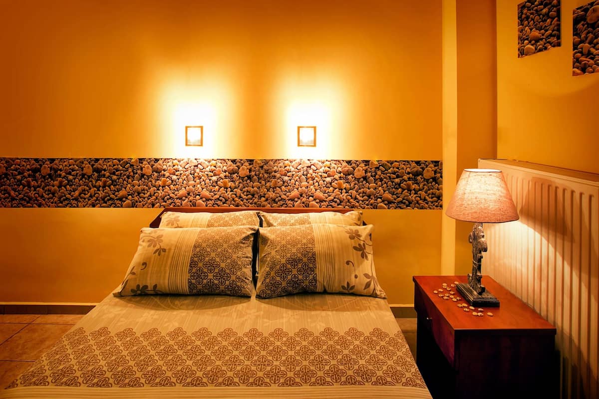 Un dormitorio ordenado con una cama, almohadas, una mesita de noche con lámpara y una pared decorada con un borde de piedrecitas.